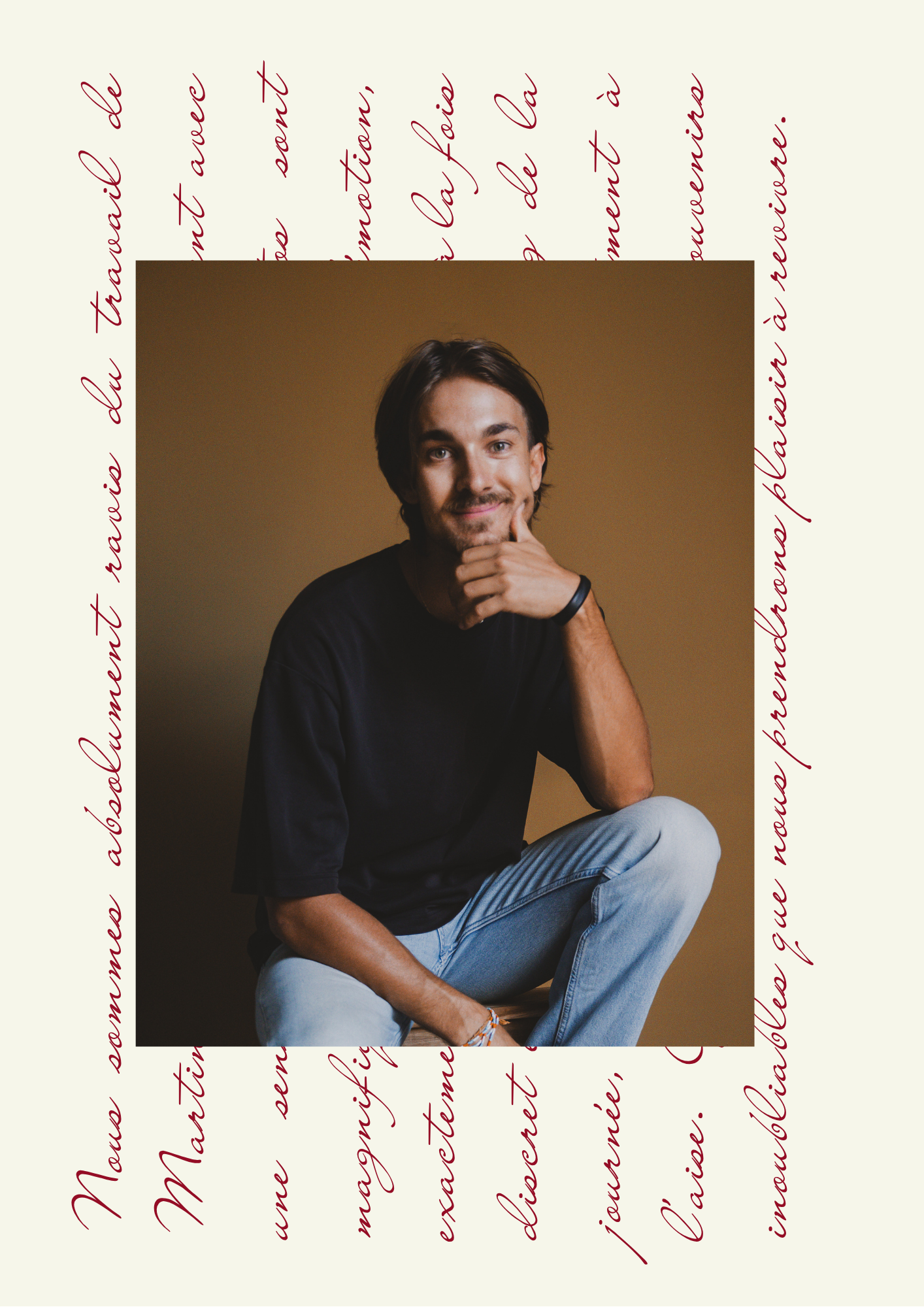 Un homme jeune, avec des yeux bleus et des cheveux bruns, assis en tailleur, portant un t-shirt noir et un bracelet noir, pose de manière détendue contre un fond beige