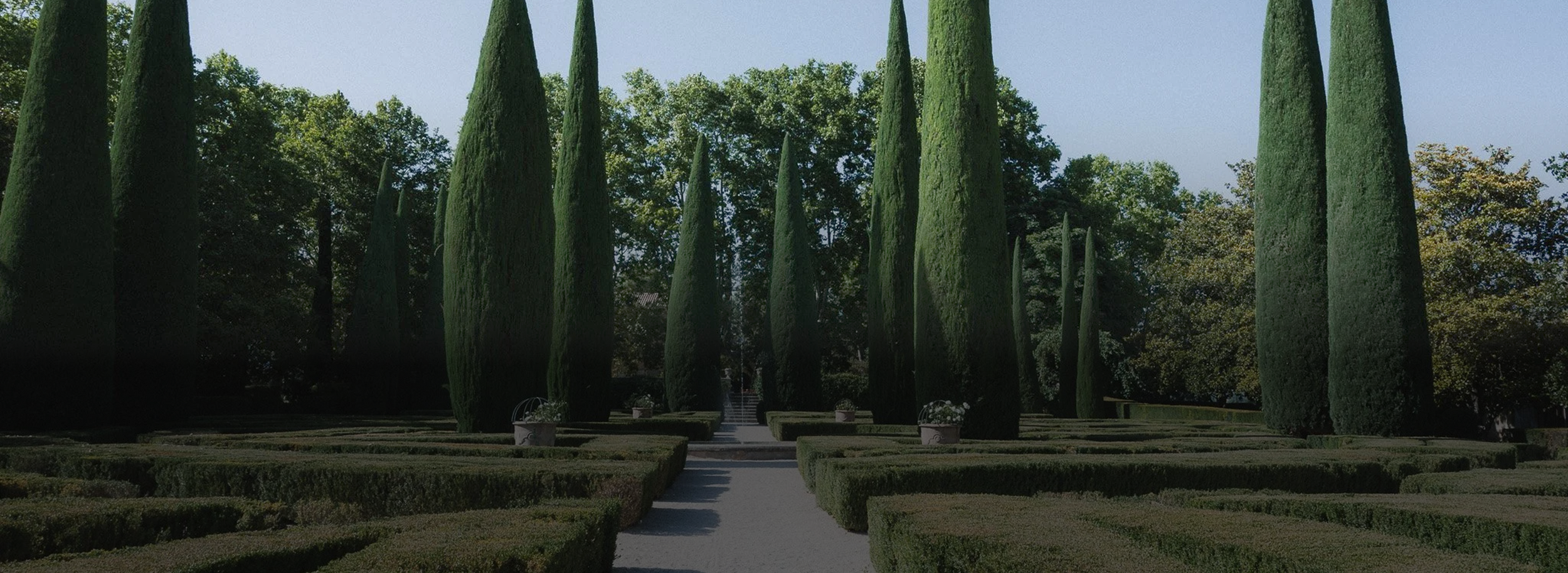 Jardin formé de hautes cyprès alignés, avec des allées rectilignes et des buissons taillés, sous un ciel clair.