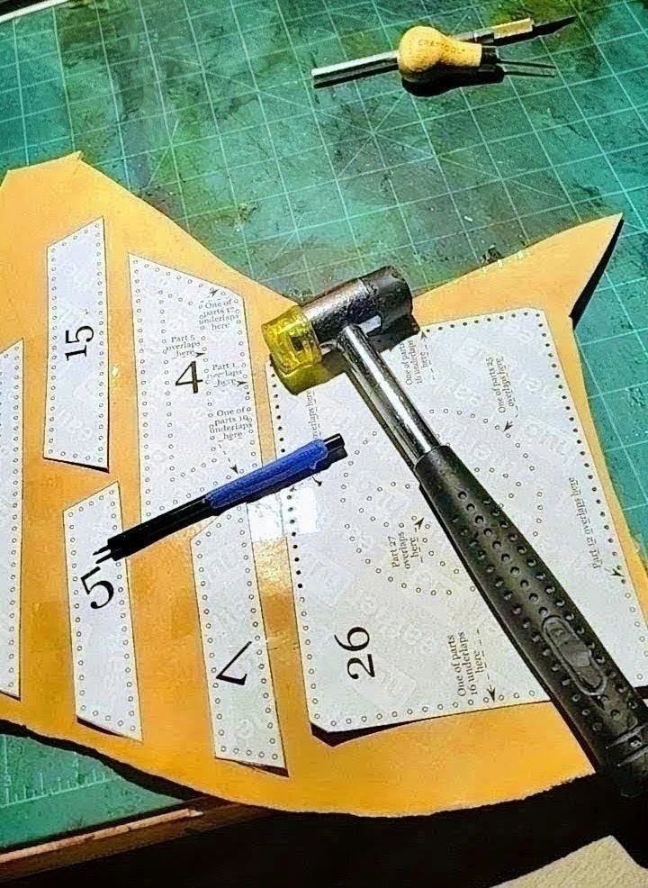 Image of a crafting workspace with a yellow foam sheet, punched-out paper pieces numbered 4, 5, 7, 15, and 26. There is a blue pen, a yellow screwdriver, and a black hammer with some foam craft materials on a green cutting mat.
