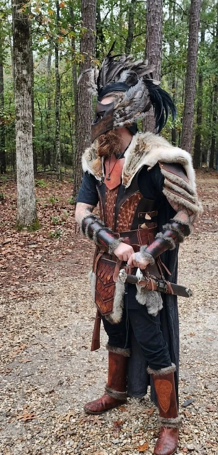 Person in medieval warrior costume with feathered helmet and fur cloak, holding a sword, standing in a forest.