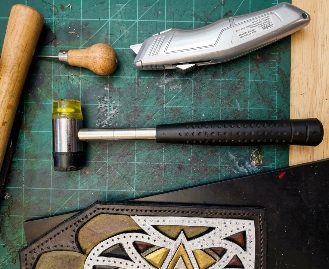 A work surface with various tools including a rubber mallet, a leather punch, an electric rotary tool, a leather sewing stitching tool, and a leather piece with decorative punched holes and a design.