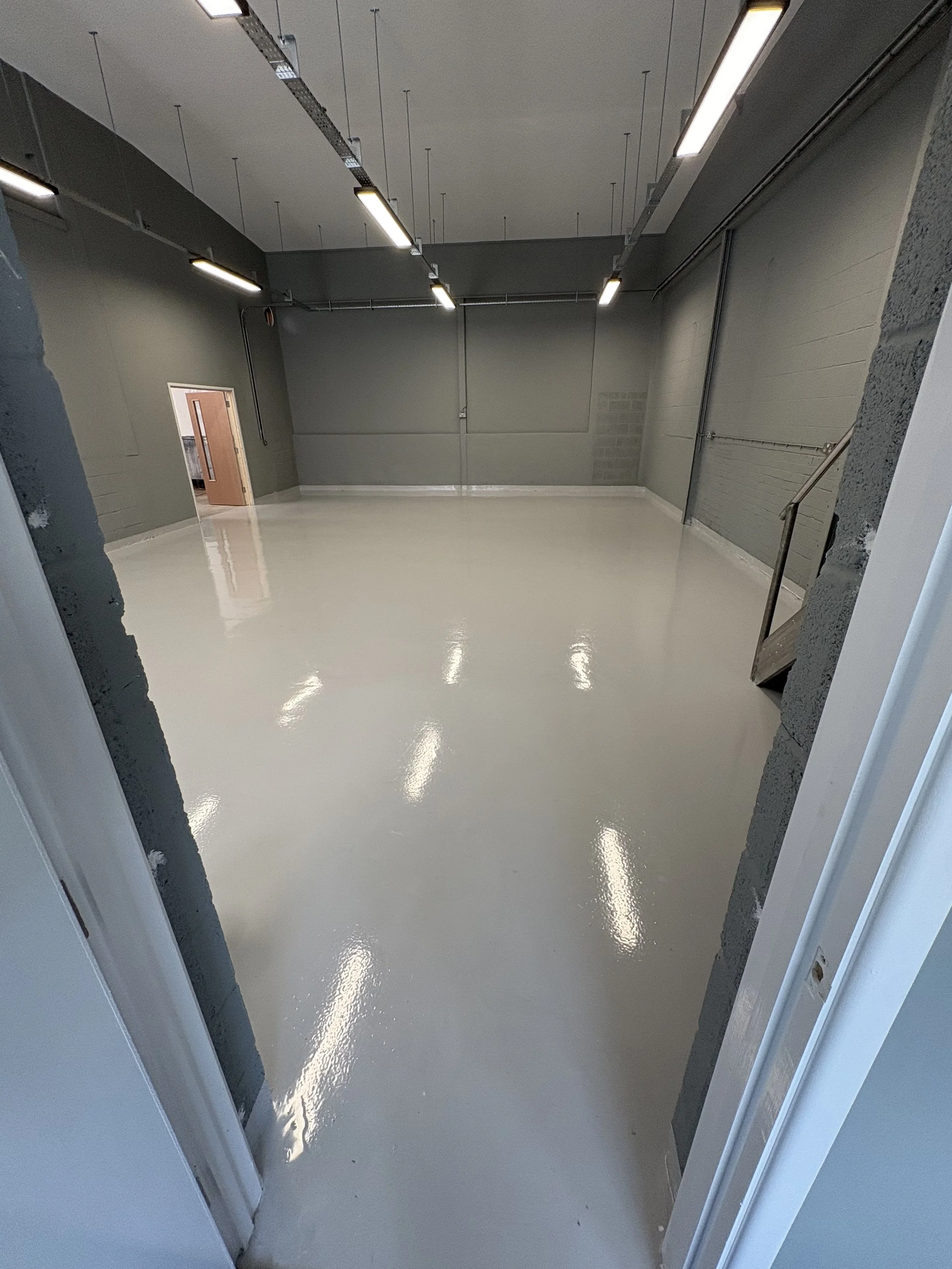 A large, empty industrial or commercial room with a shiny, polished concrete floor, gray walls, and fluorescent lighting fixtures hanging from the ceiling. There is a small door on the left wall and an open doorway in the foreground, showing part of a staircase to the right.