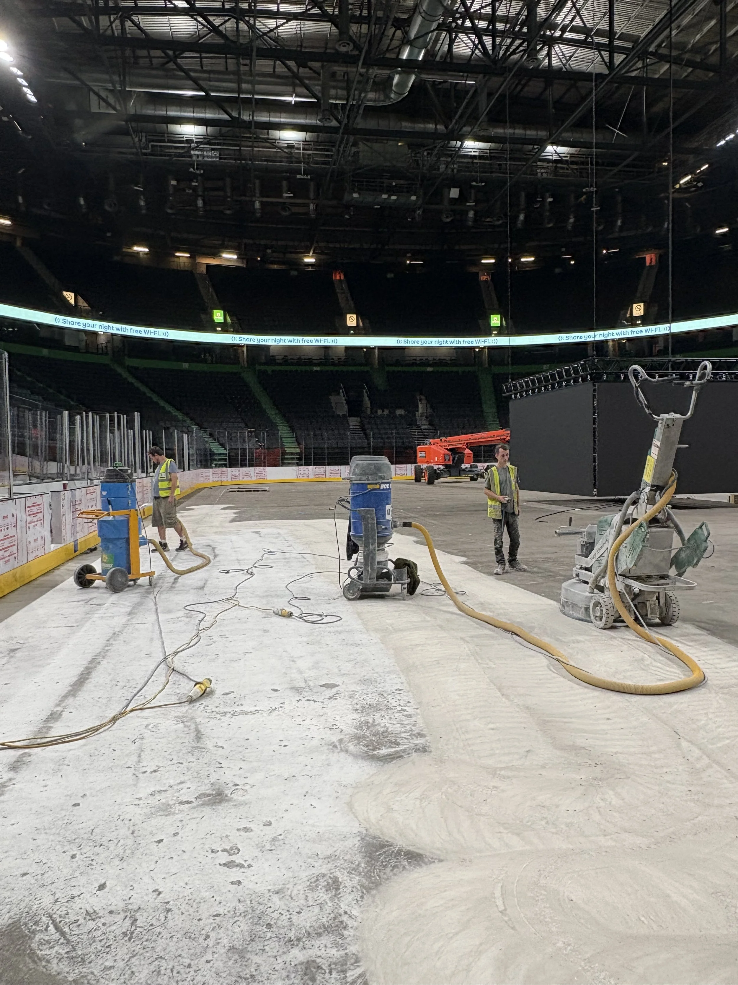 An indoor hockey arena undergoing renovation or cleaning, with workers in safety vests and construction equipment scattered across the ice surface.