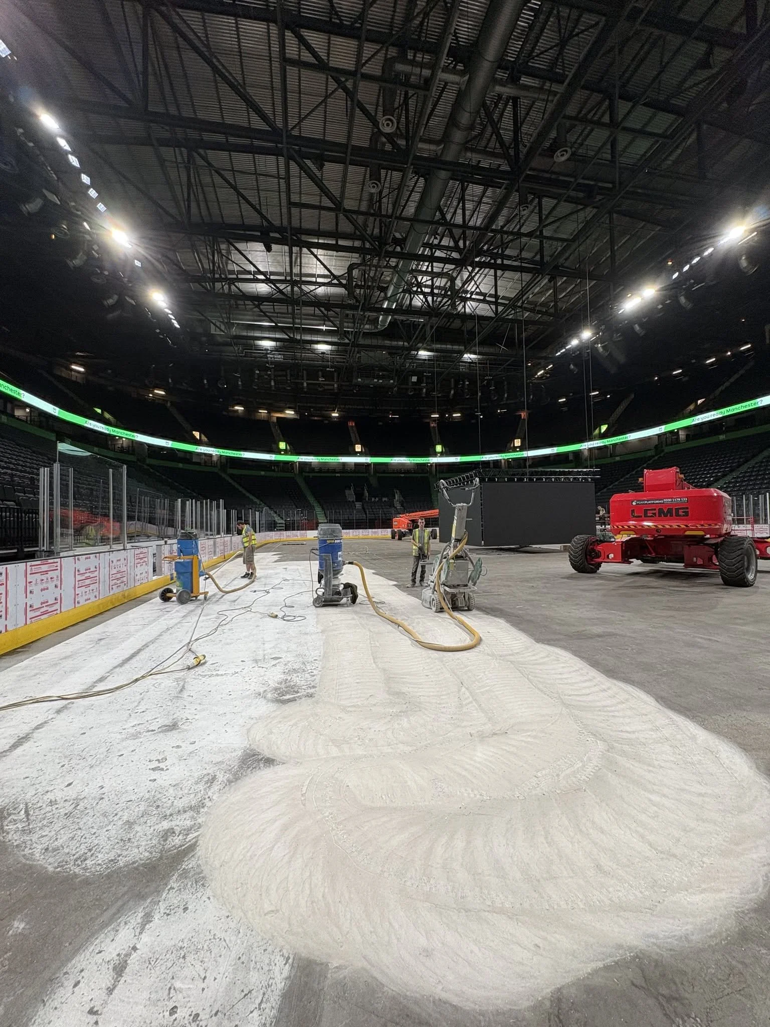 Inside an ice hockey arena with workers installing or repairing the ice rink surface. Construction equipment and materials are visible, including a red lift, hoses, and power tools. The arena is well-lit with overhead lighting and has seating around the rink.