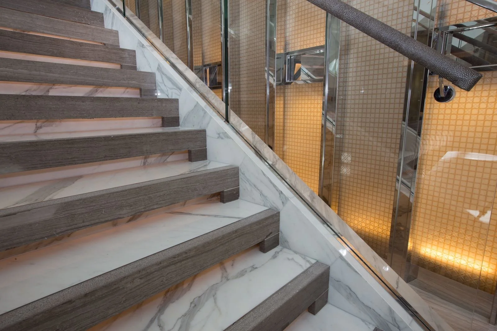 Modern marble staircase with dark wood treads, glass railing, and illuminated golden tile walls