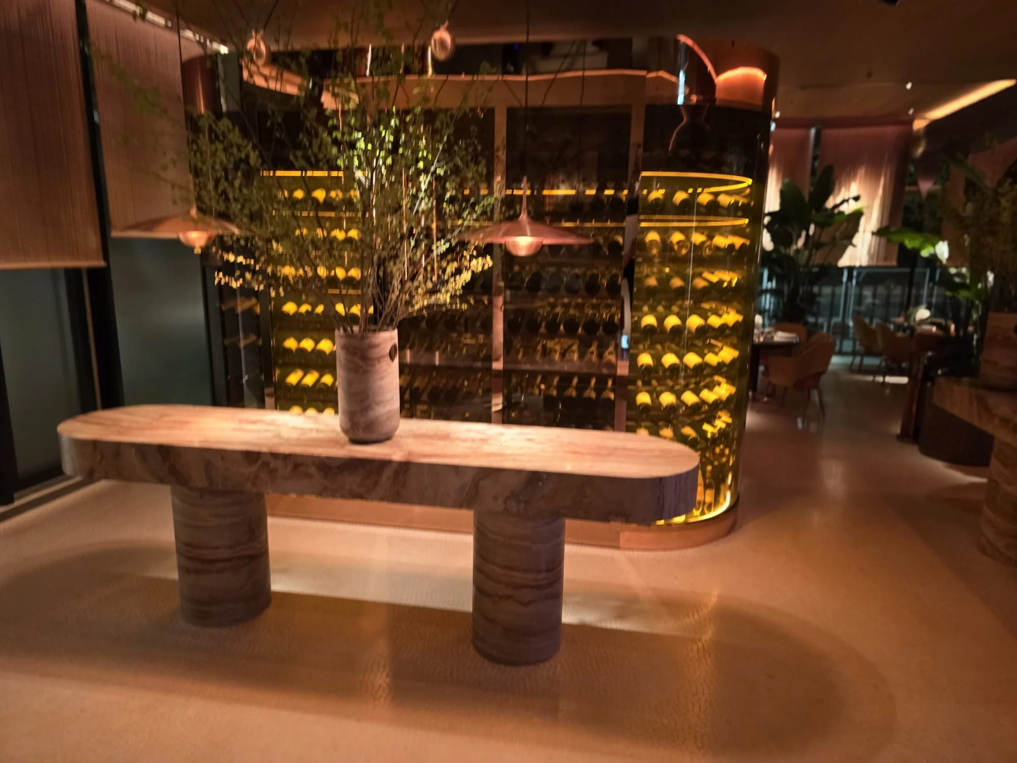 Warmly lit restaurant wine cellar with glass racks of wine bottles, a curved stone console table, and a potted plant.