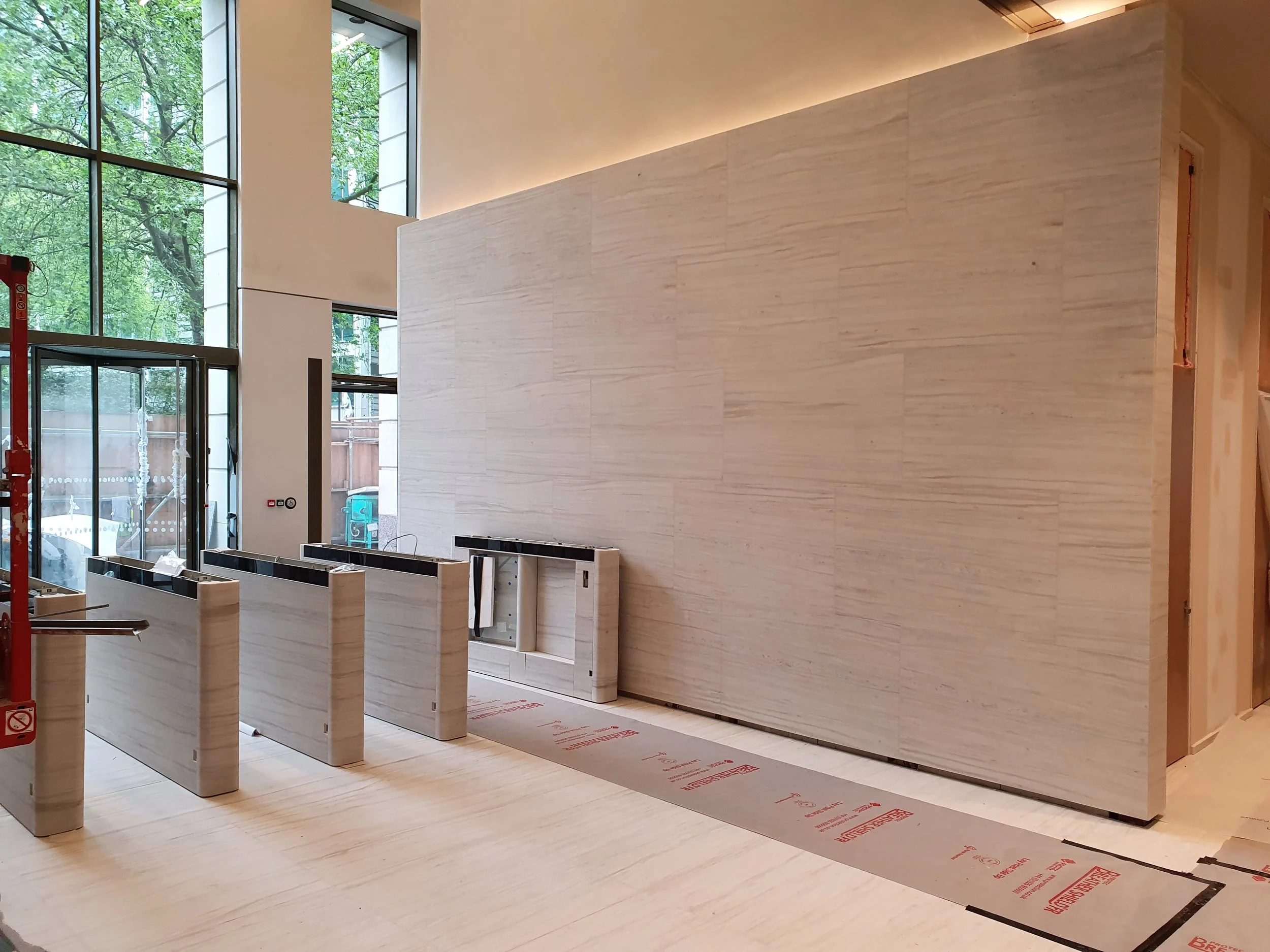 Modern building lobby under construction with pale stone reception counters, a large textured stone wall, floor protection, and floor-to-ceiling windows.