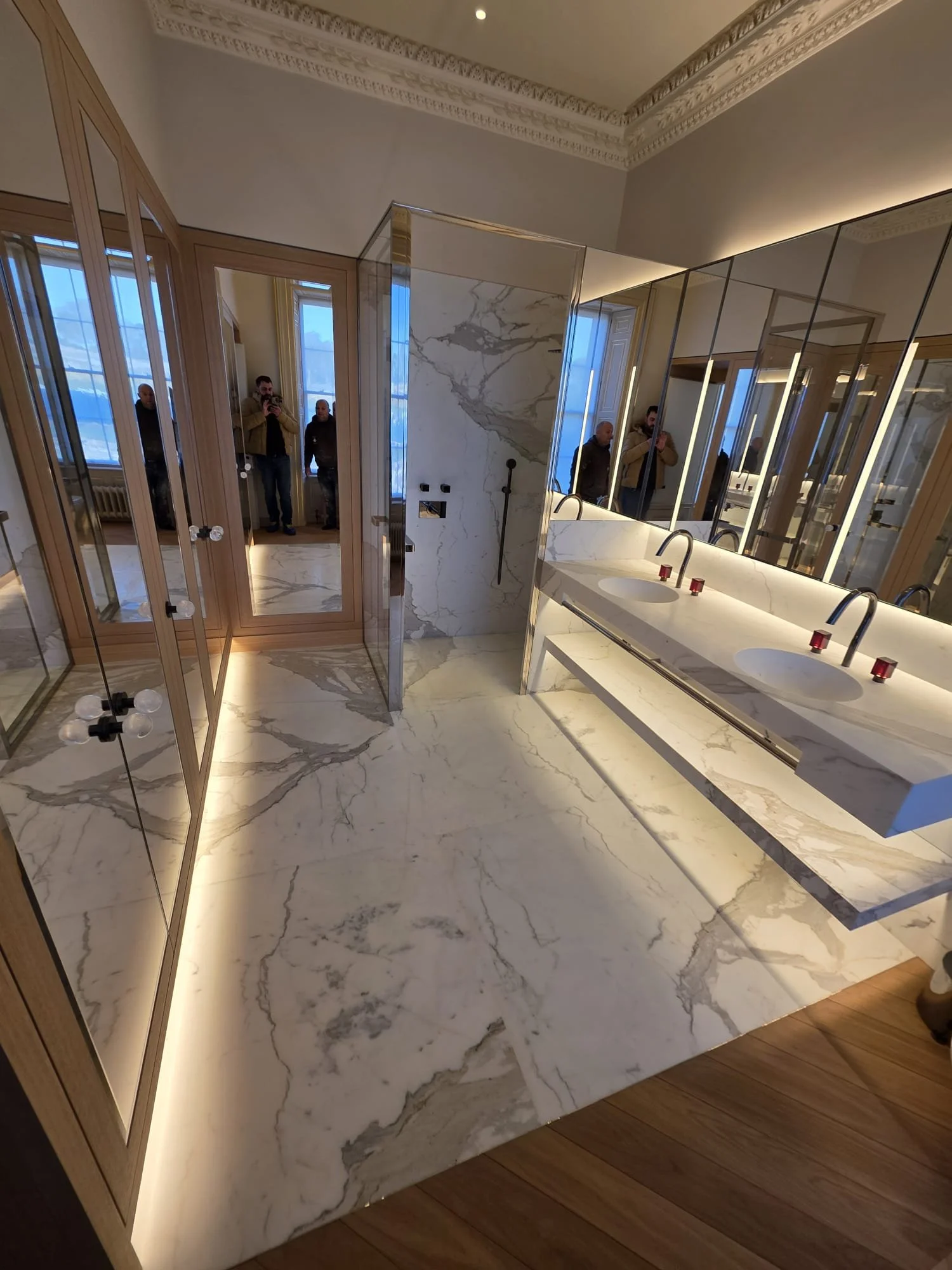 Luxury marble bathroom with double vanity sinks, illuminated mirrors, glass shower enclosure, and decorative molding