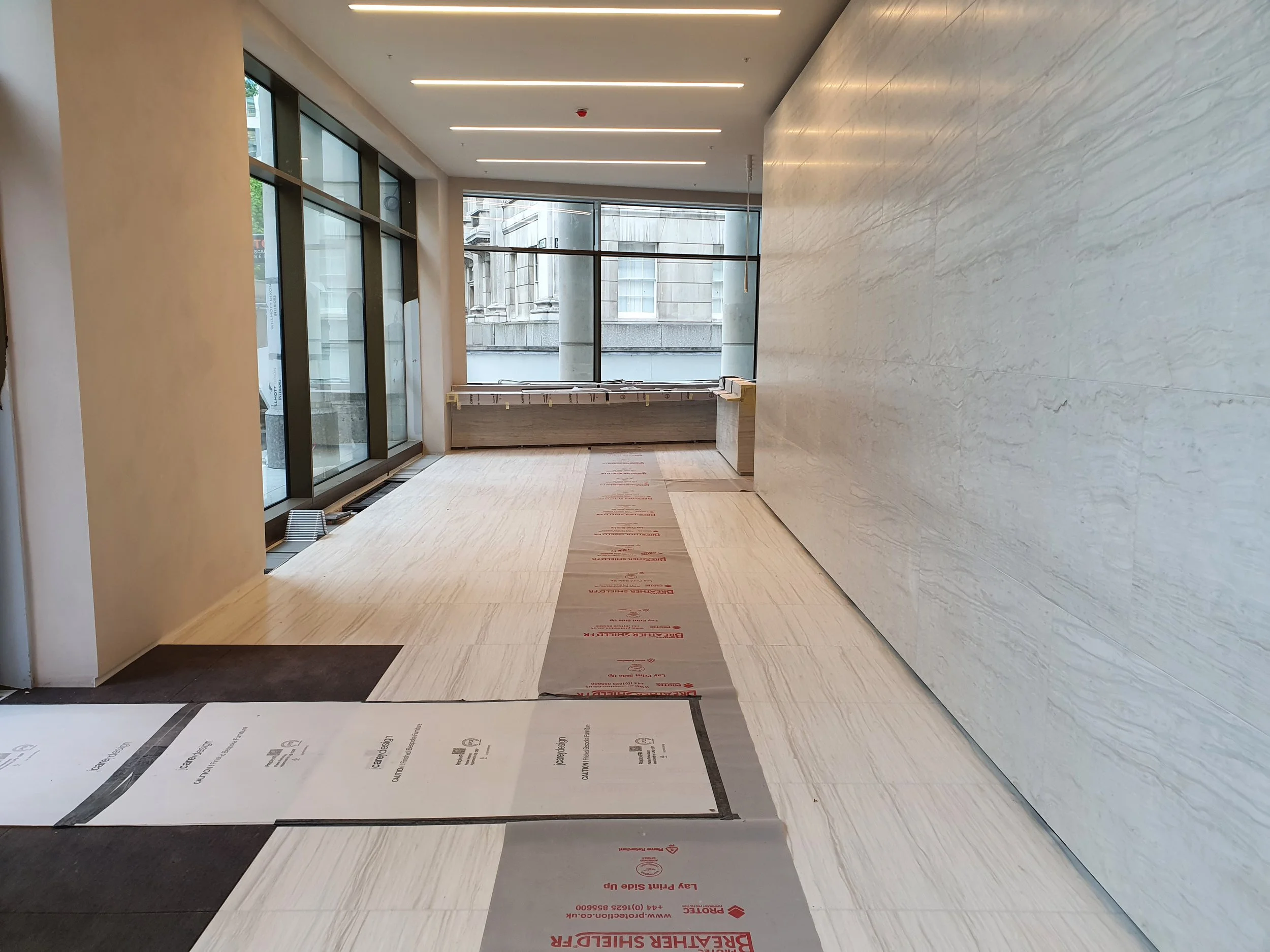 Modern building lobby under construction with protective floor coverings, marble walls, large windows, and recessed linear lighting