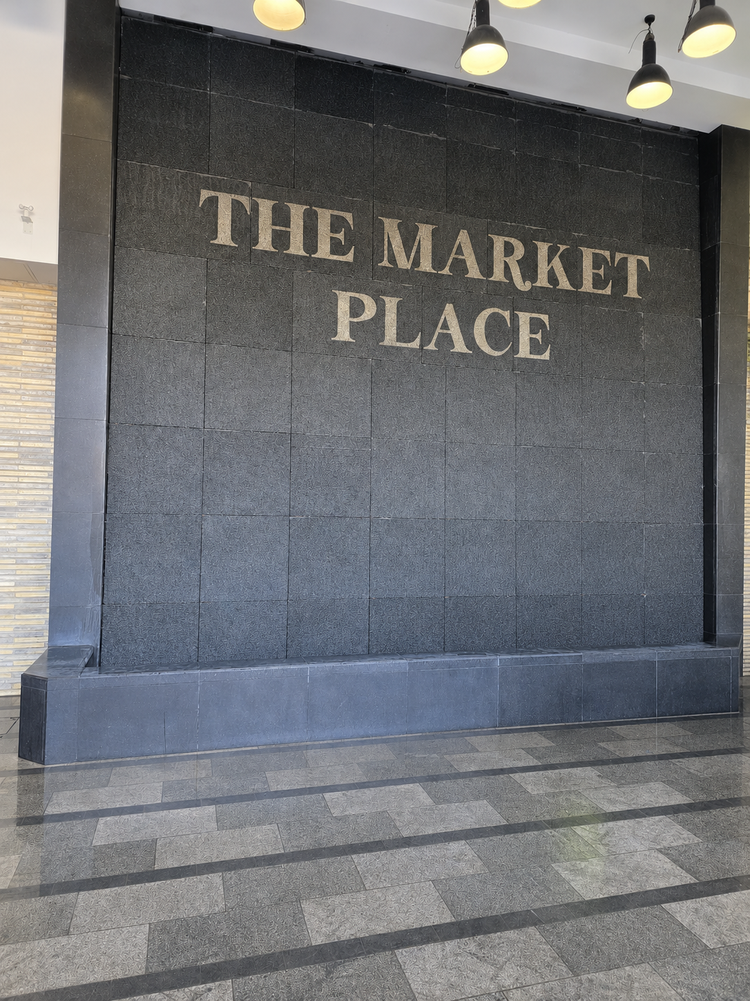 Interior wall with large lettering reading “THE MARKET PLACE,” black tile panels, pendant lights, and patterned stone floor.