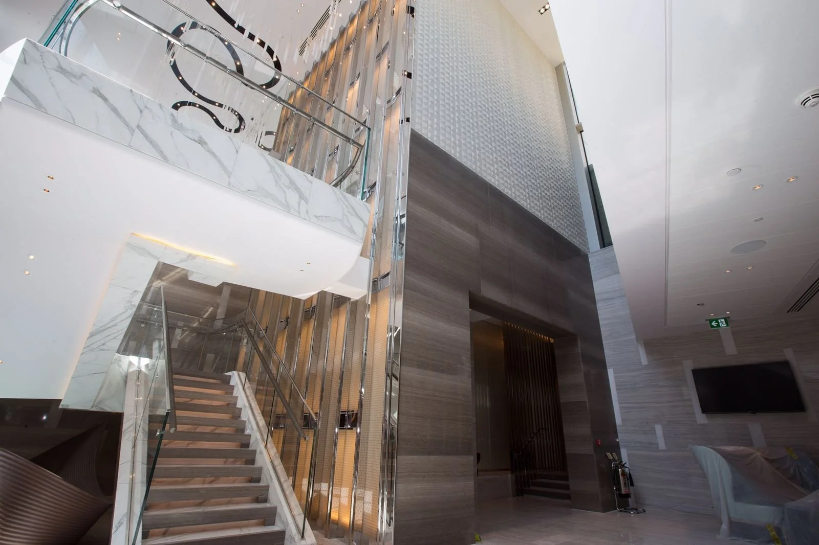 Modern luxury interior lobby with marble staircase, glass railings, elevator, wood-paneled walls, and ceiling lighting