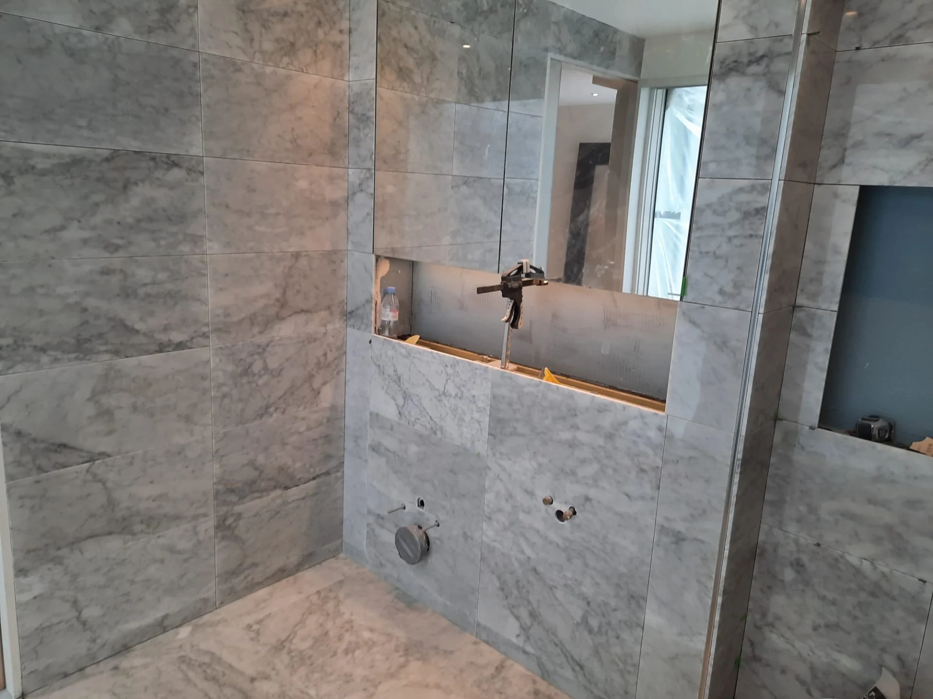 Unfinished bathroom with gray marble-look tile, a recessed medicine cabinet opening, exposed plumbing, and construction tools.
