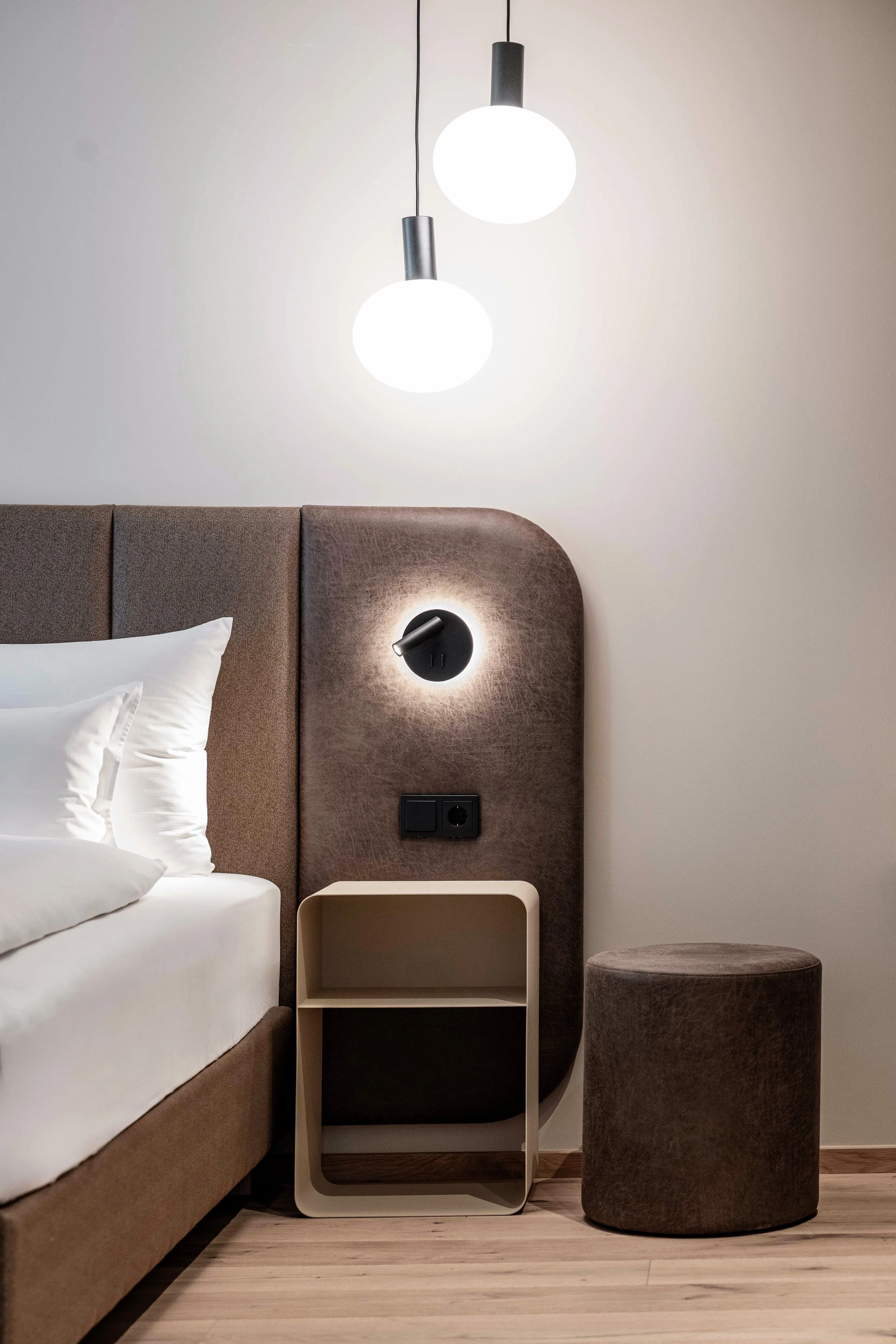Ein modernes Schlafzimmer mit einem braunen Bett, weißen Bettwäsche, zwei weißen Kissen, Nachttisch, Wandlampe, runden schwarzen Licht, Stehlampe, und grauem Pouf, mit hellem Holzfußboden.