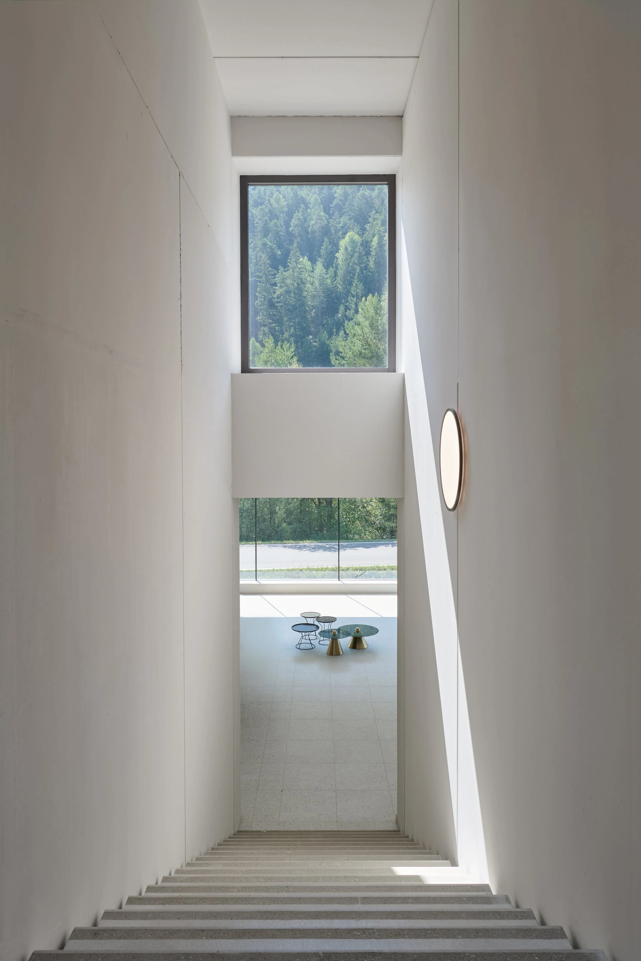 Innenansicht mit weißen Wänden, Treppe und großem Fenster mit Blick auf Bäume und Natur, moderne minimalistische Gestaltung mit runden Tischen und Stühlen