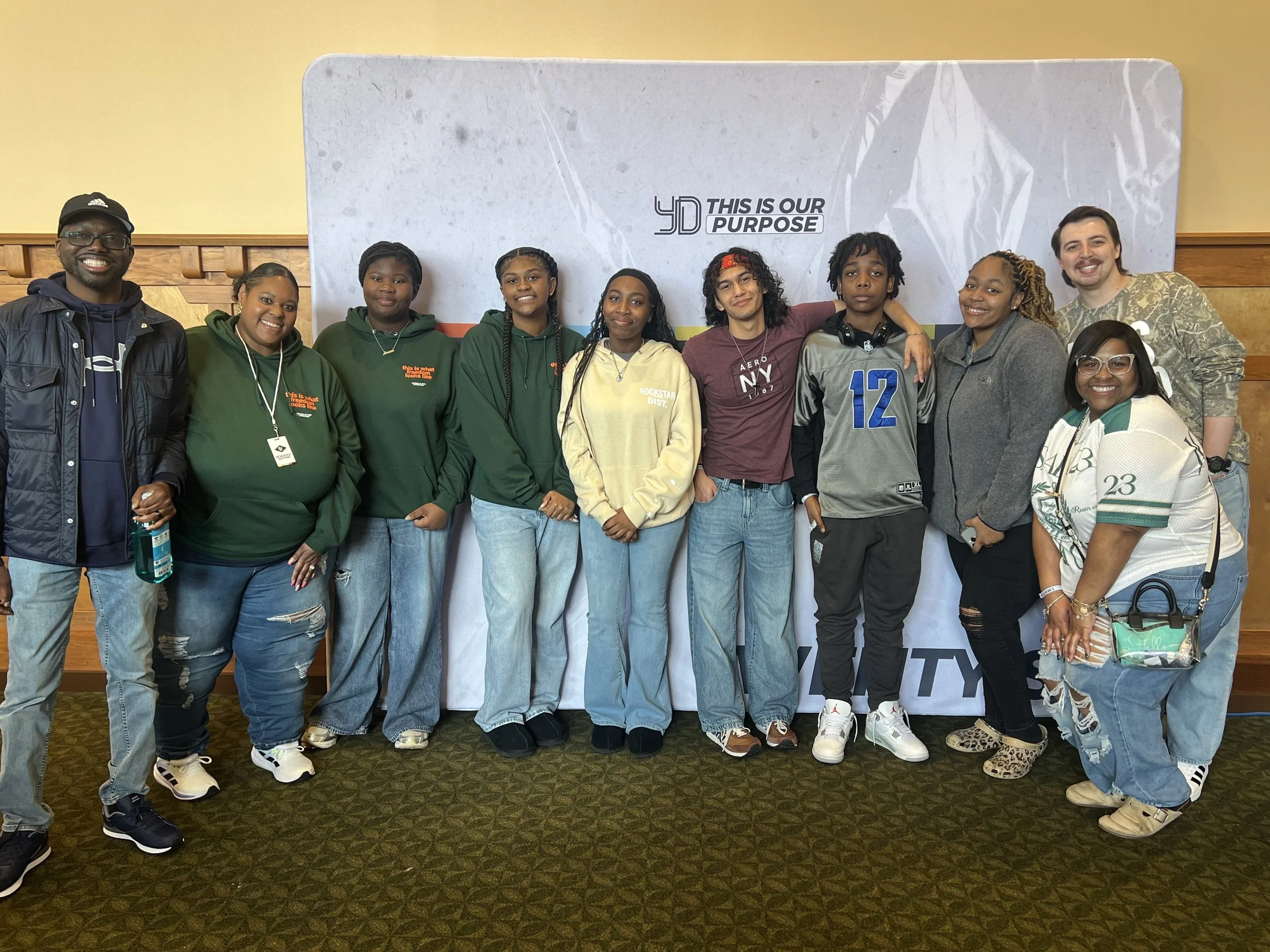 A group of ten diverse people, including men and women, standing together indoors in front of a large backdrop that reads '4D This is Our Purpose.' They are smiling and dressed casually, with some wearing hoodies, jackets, and sneakers.