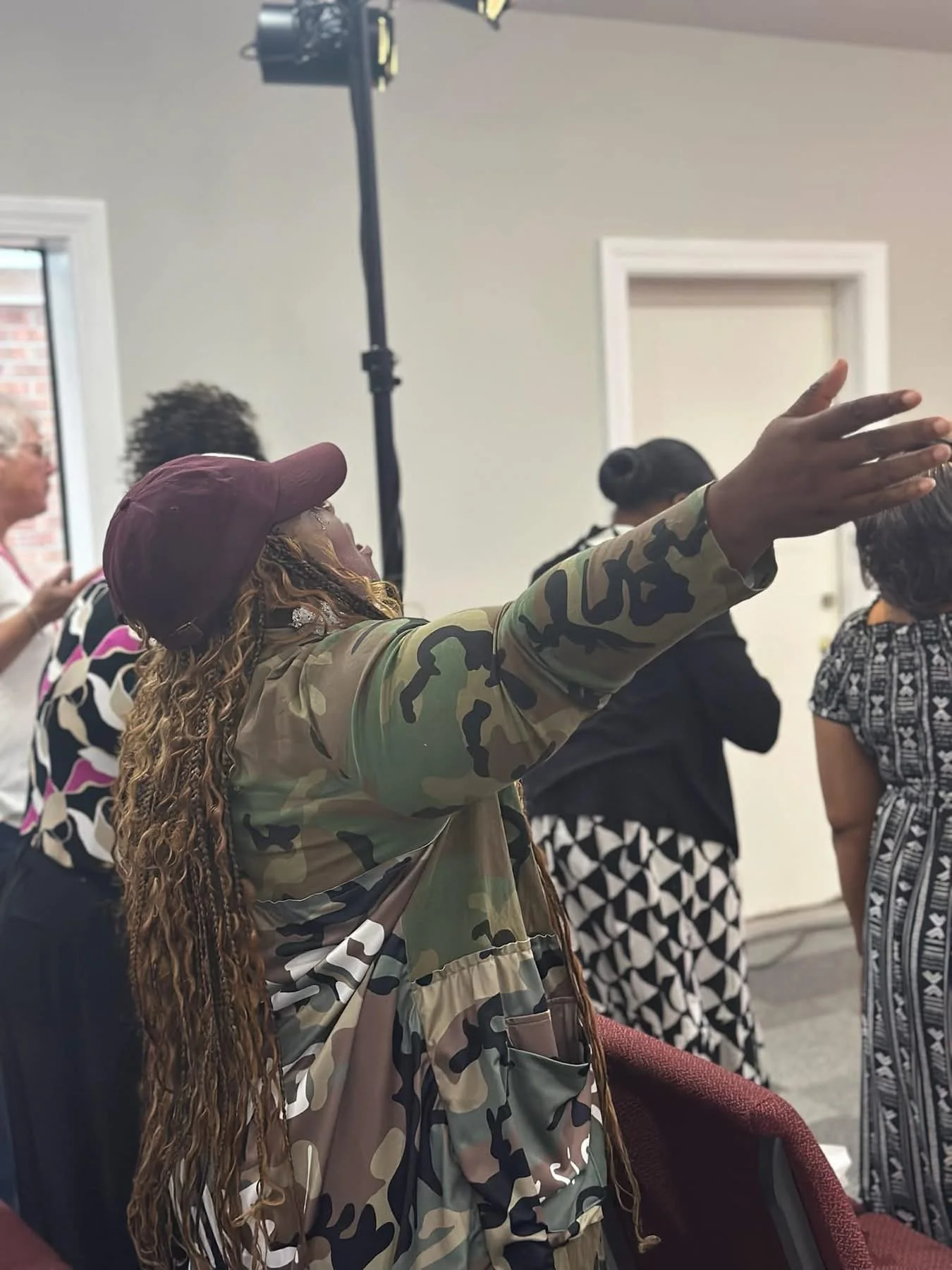 A group of women standing indoors, one woman with long, curly hair and a camouflage jacket is raising her hand, wearing a cap. Others are in patterned dresses or shirts, standing around in a room with light-colored walls and a window.