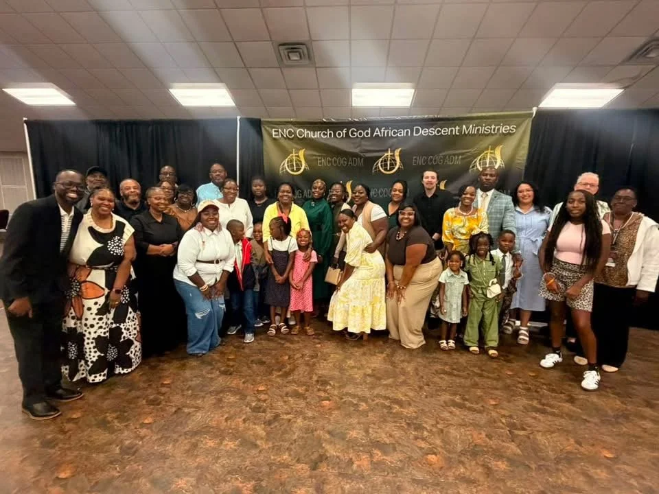 Group photo of diverse adults and children standing in front of a banner that reads 'ENC Church of God African Descent Ministries' inside a community hall.