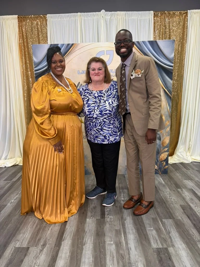 Three people standing in front of a decorated backdrop, smiling for a photo. The woman on the left is wearing a gold pleated gown, the woman in the middle is wearing a blue and white patterned shirt with black pants, and the man on the right is weari