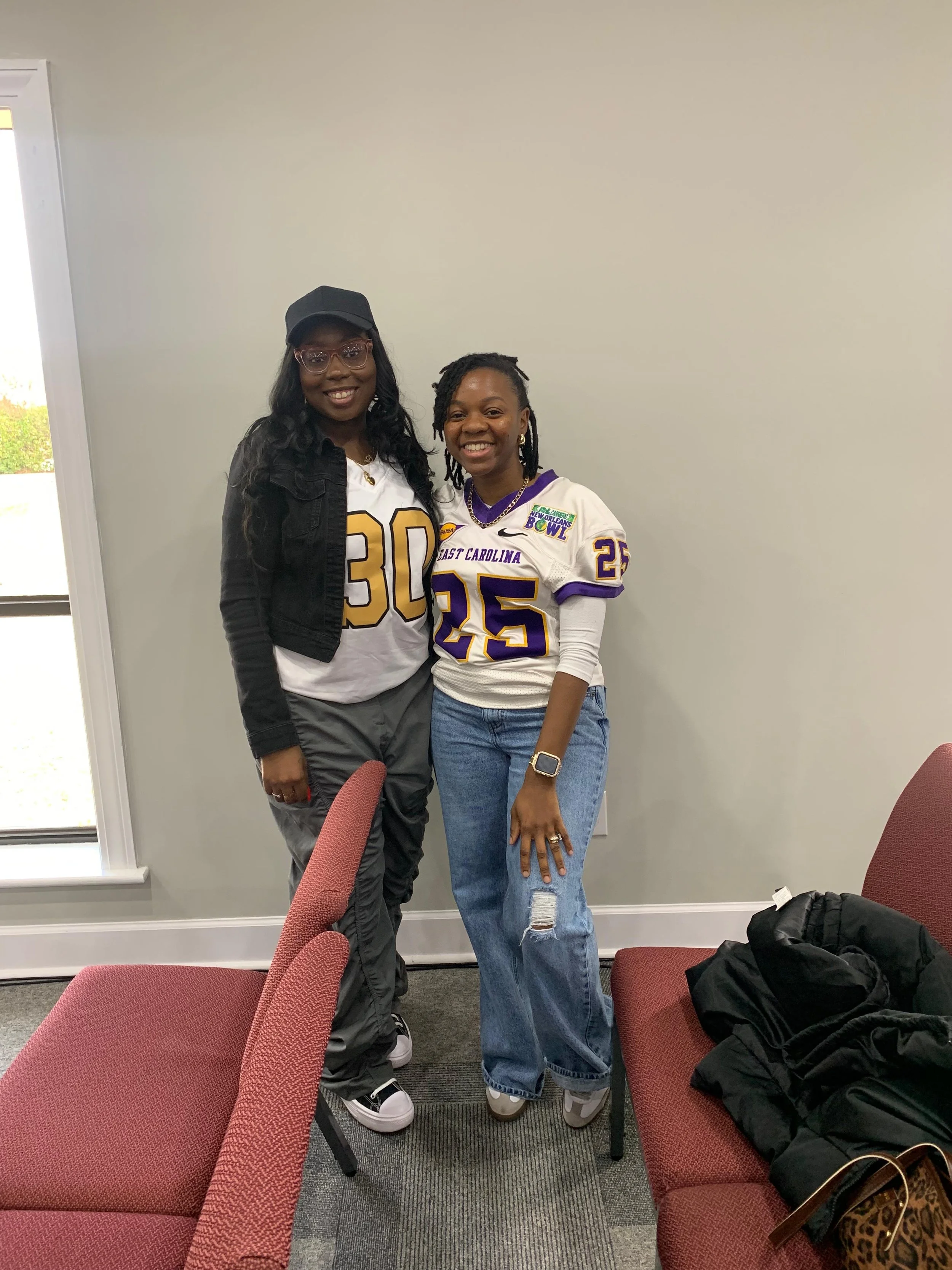 Two women smiling and posing for a photo indoors, one wearing a black jacket and cap, the other wearing a white football jersey with purple and gold details, sitting on red chairs, with a window and plain wall in the background.