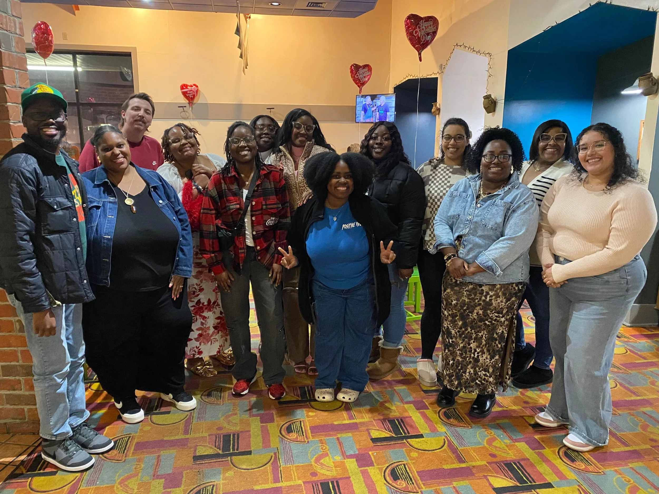 A diverse group of people posing together indoors at a celebration, with Valentine's Day balloons and festive decorations in the background.