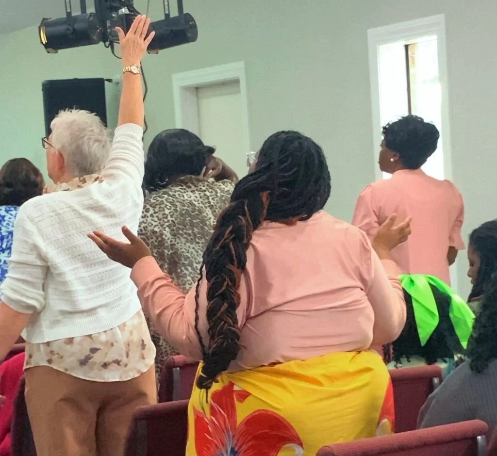 Group of women standing in a room, some raising their hands or gesturing, with a green wall and windows in the background.