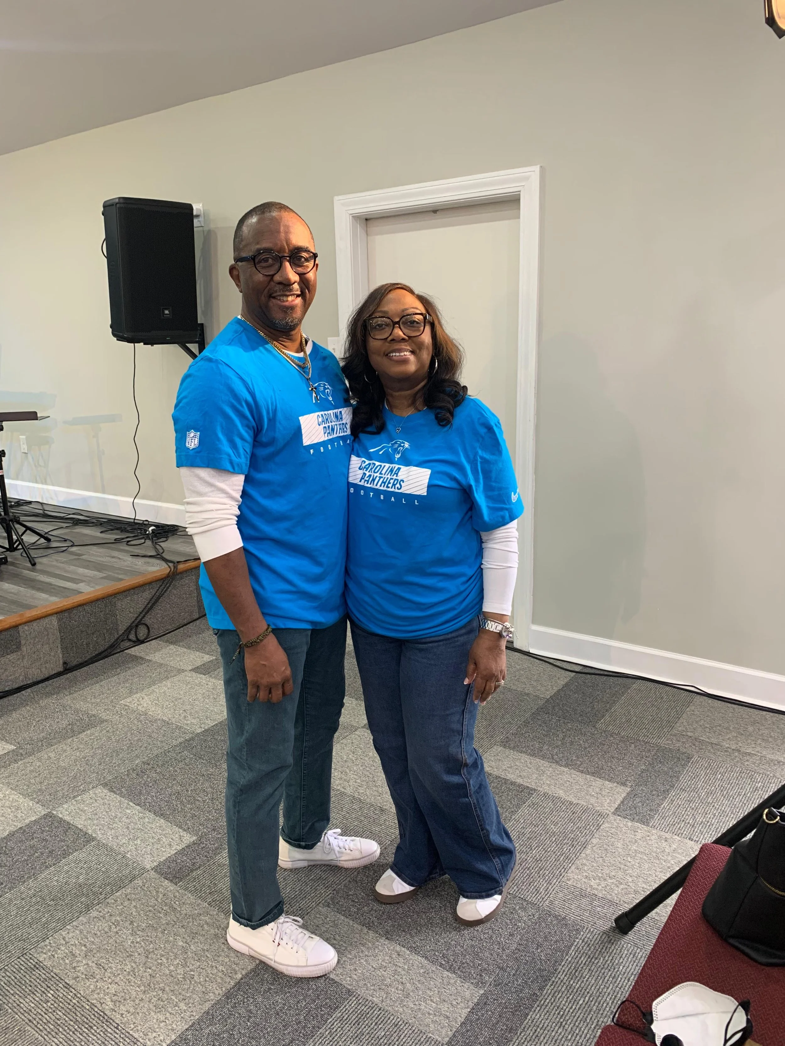 A man and a woman standing together indoors, both wearing blue Carolina Panthers football shirts and smiling at the camera.
