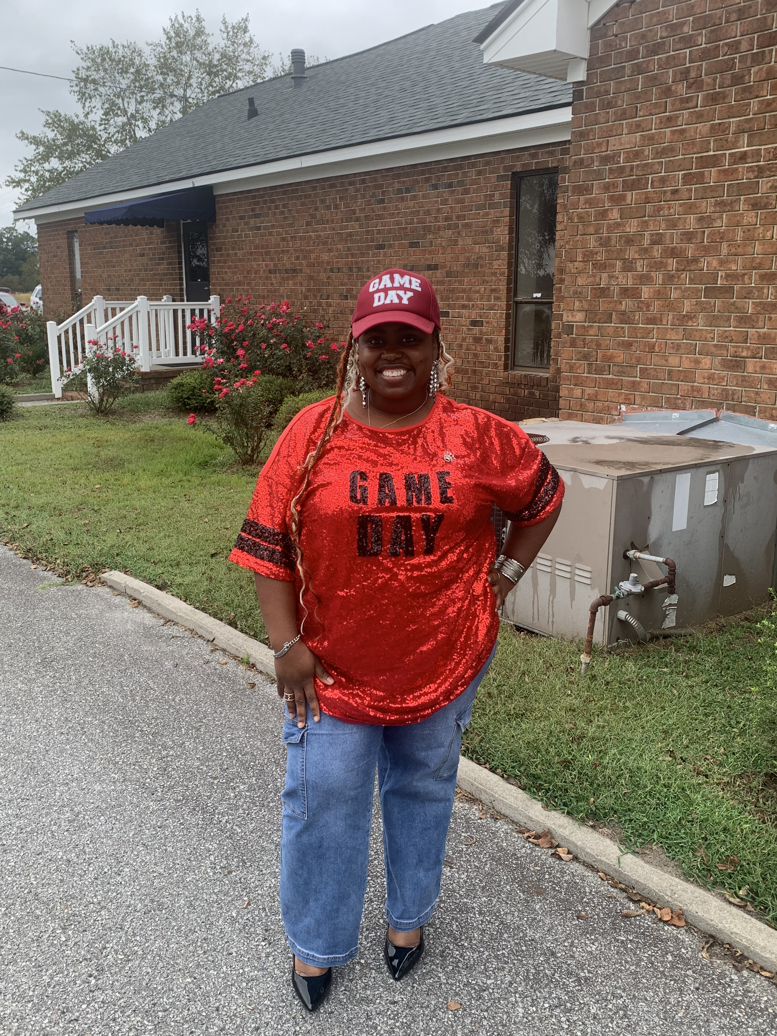 A woman standing outside of lion of Judah community church, wearing a red shirt and a red cap that says "GAME DAY," and blue jeans. She is smiling and posing for the photo.