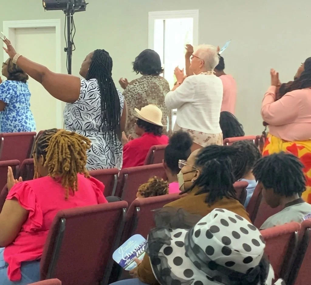 A diverse group of people standing and sitting in a church or community hall, some with hands raised in prayer or praise, participating in a service or gathering.