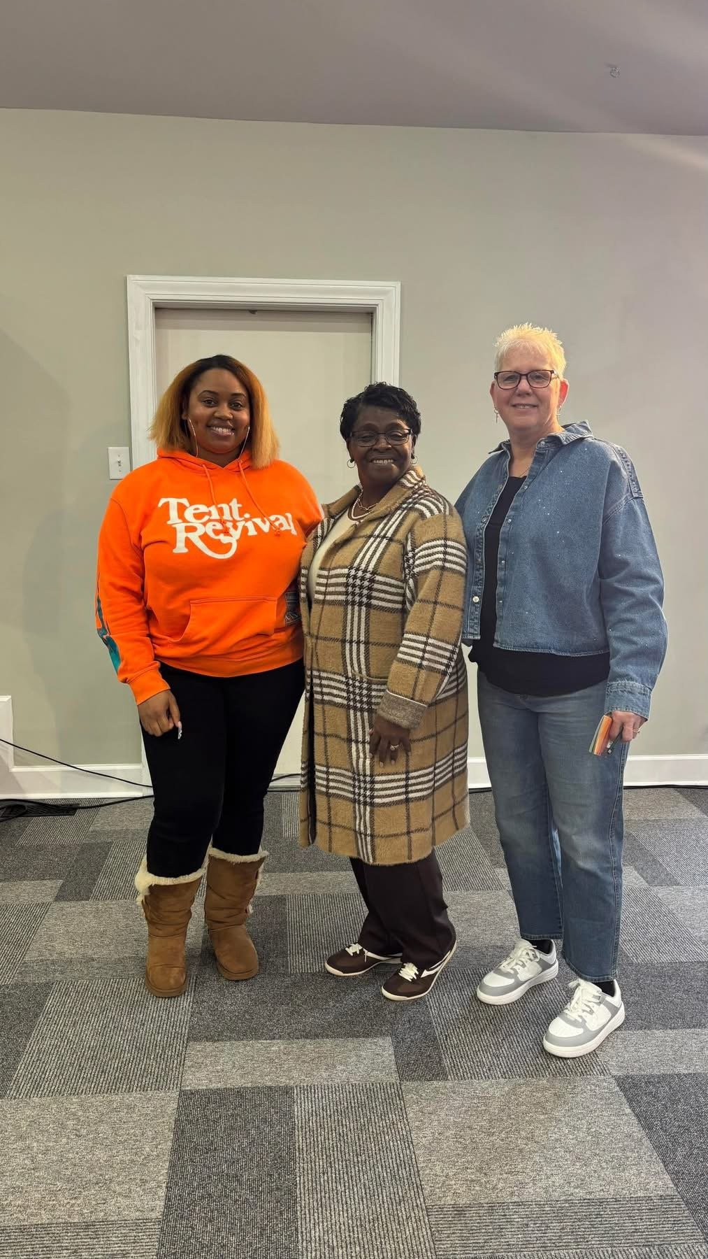 Three women standing together indoors in front of a plain wall with a white door, smiling at the camera.