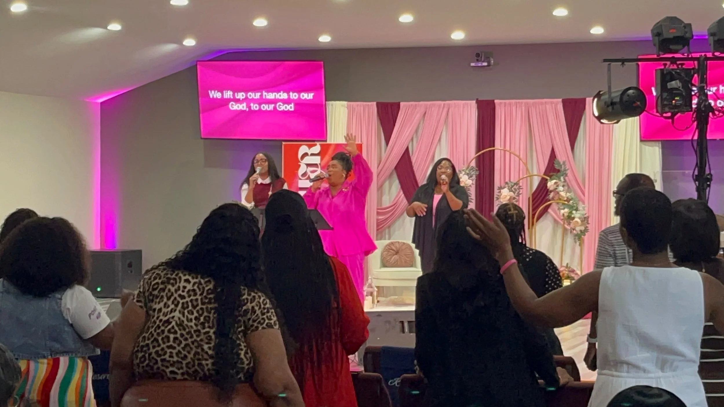 Indoor church service with women singing and raising hands, pink and purple decorative backdrop, congregation in attendance, LED screens displaying lyrics about worshiping God.