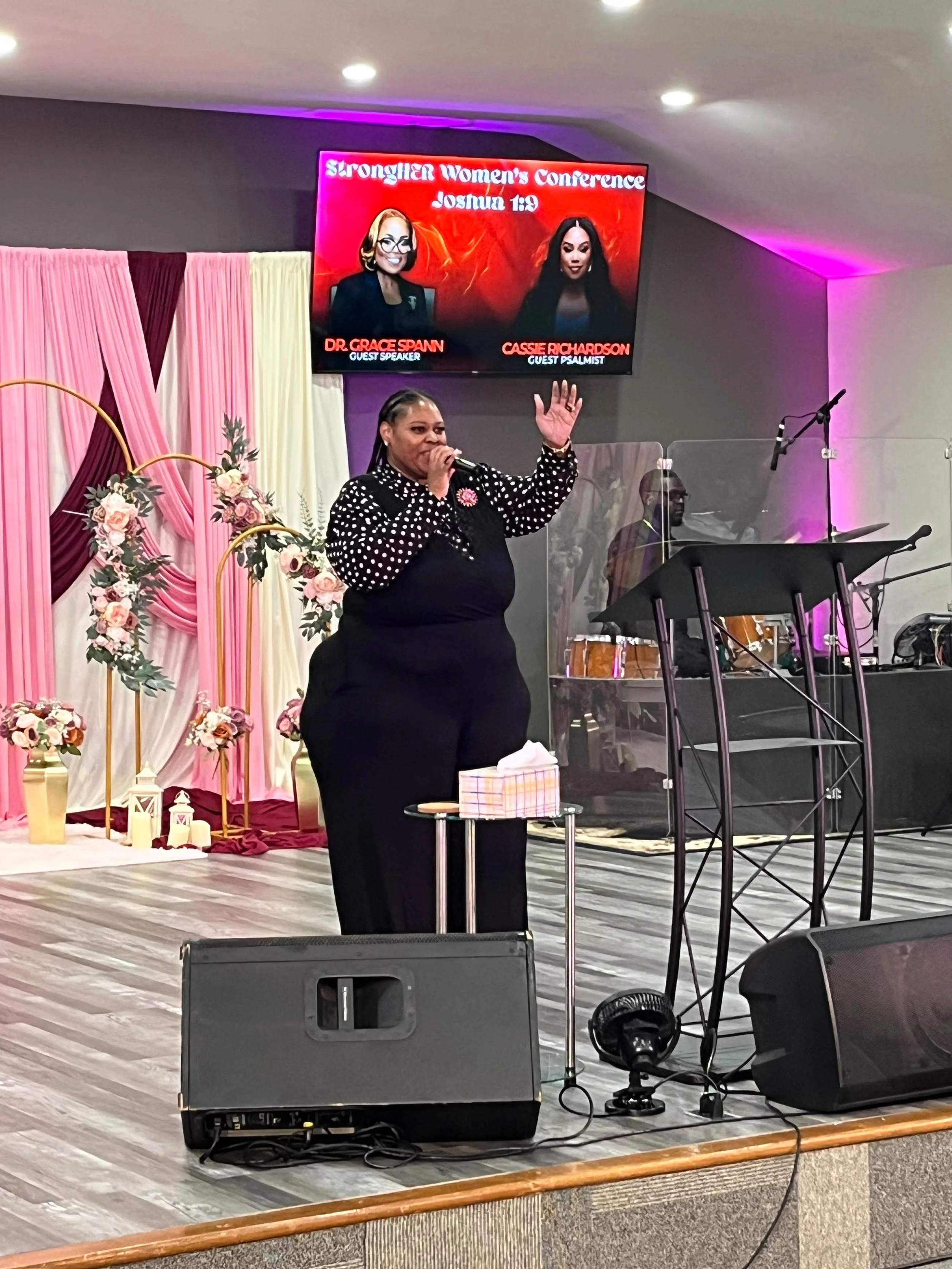 Woman speaking into a microphone on a stage decorated with pink and cream drapes and floral arrangements, during a women's conference. A large screen behind her displays the event's title and speaker information.
