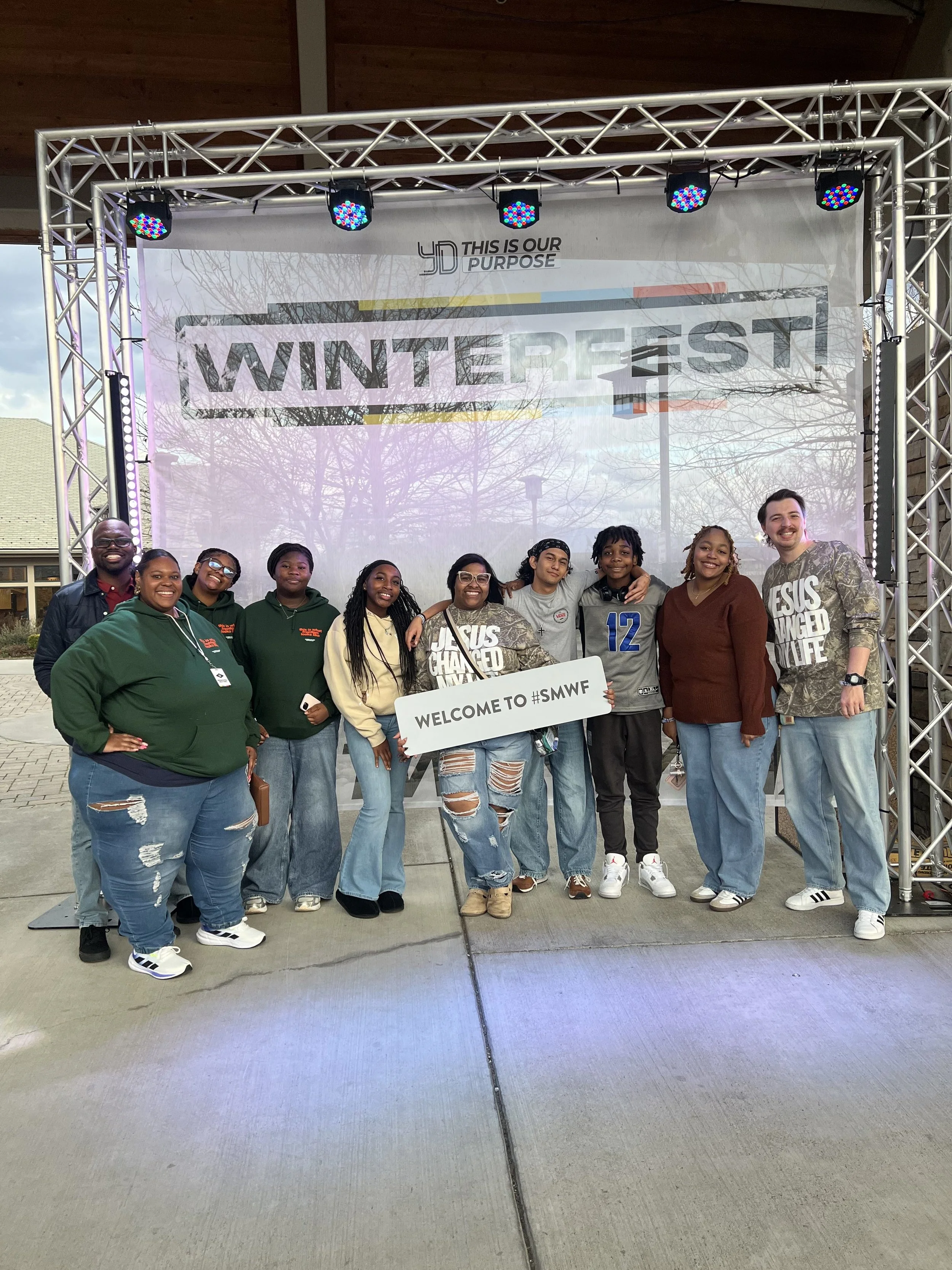 A group of people standing in front of a large display that says 'Winter Fest' and a sign that says 'Welcome to #SMWF'. They are smiling and posing for the photo, with some holding a sign. The backdrop has a tree reflection and a partial view of a bu
