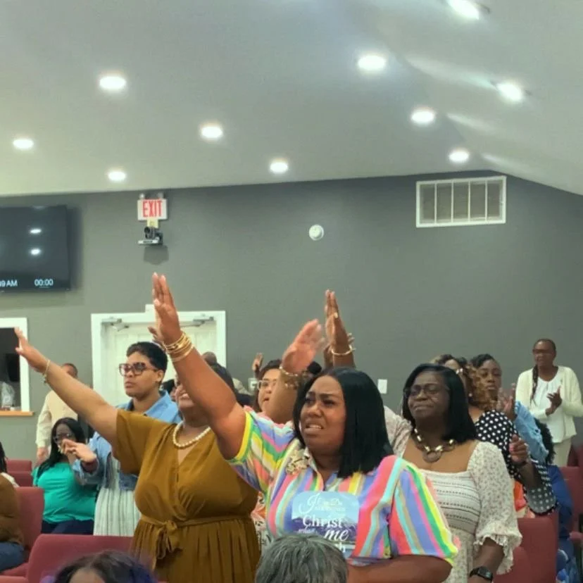Group of women and men at a church service, some with raised hands, in a room with gray walls and ceiling lights.