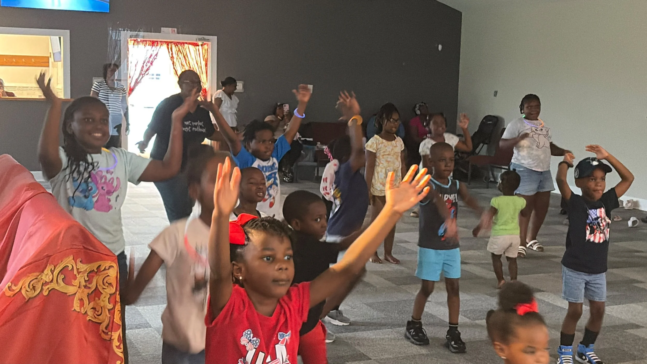 Judah Kids and adults of Lion of Judah Community Church dancing and raising their hands in a room with a gray and white wall and curtains in the background.