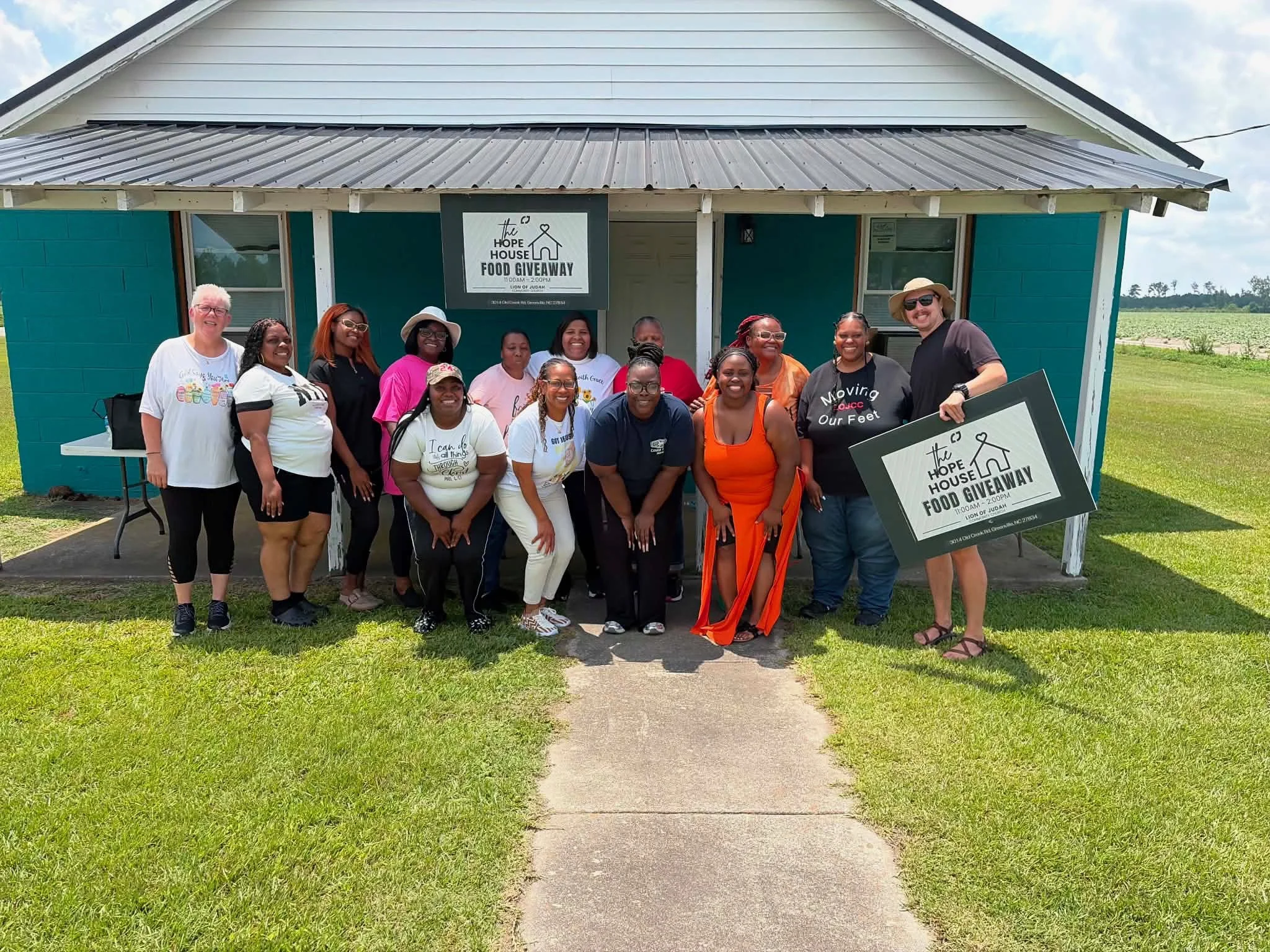 Members of Lion of Judah Community Church standing in front of Judah Hope House a small teal house with a silver roof, smiling for a photo during a food giveaway event.