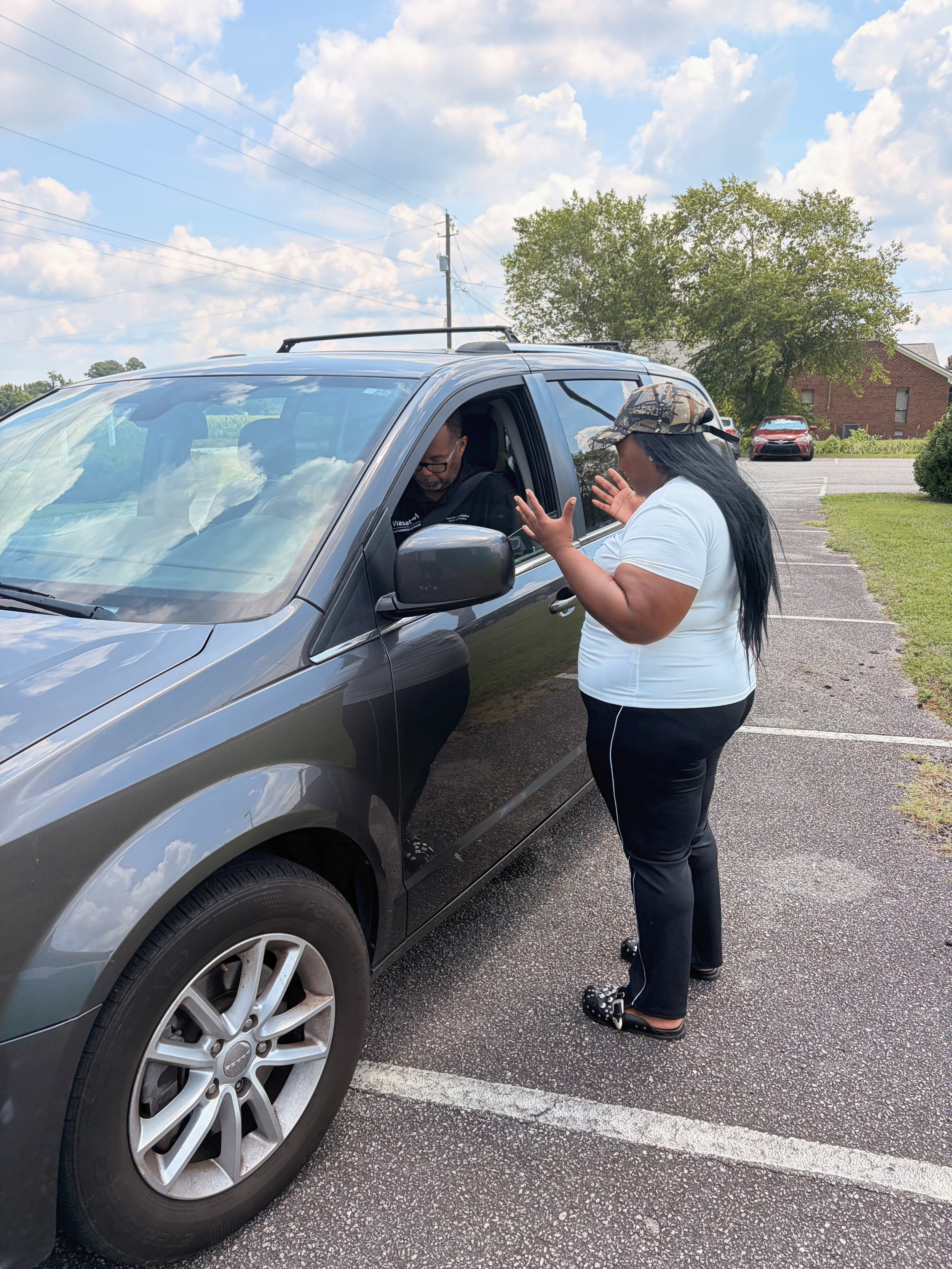 A woman is talking to a man in a car in a parking lot on a sunny day. The woman is wearing a camouflage cap, a white shirt, and black pants with white stripes. The man is inside the vehicle, wearing glasses and a black hoodie. The parking lot has multiple cars and is next to a grassy area and trees, with a building and electrical wires in the background.