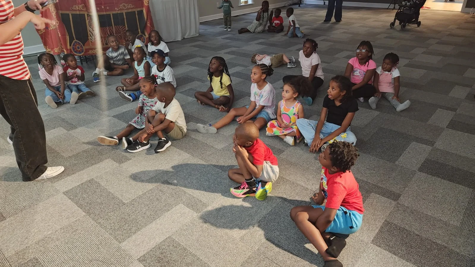 A group of children sitting on the carpeted floor, attentively looking at a person pointing and giving a presentation or instructions.