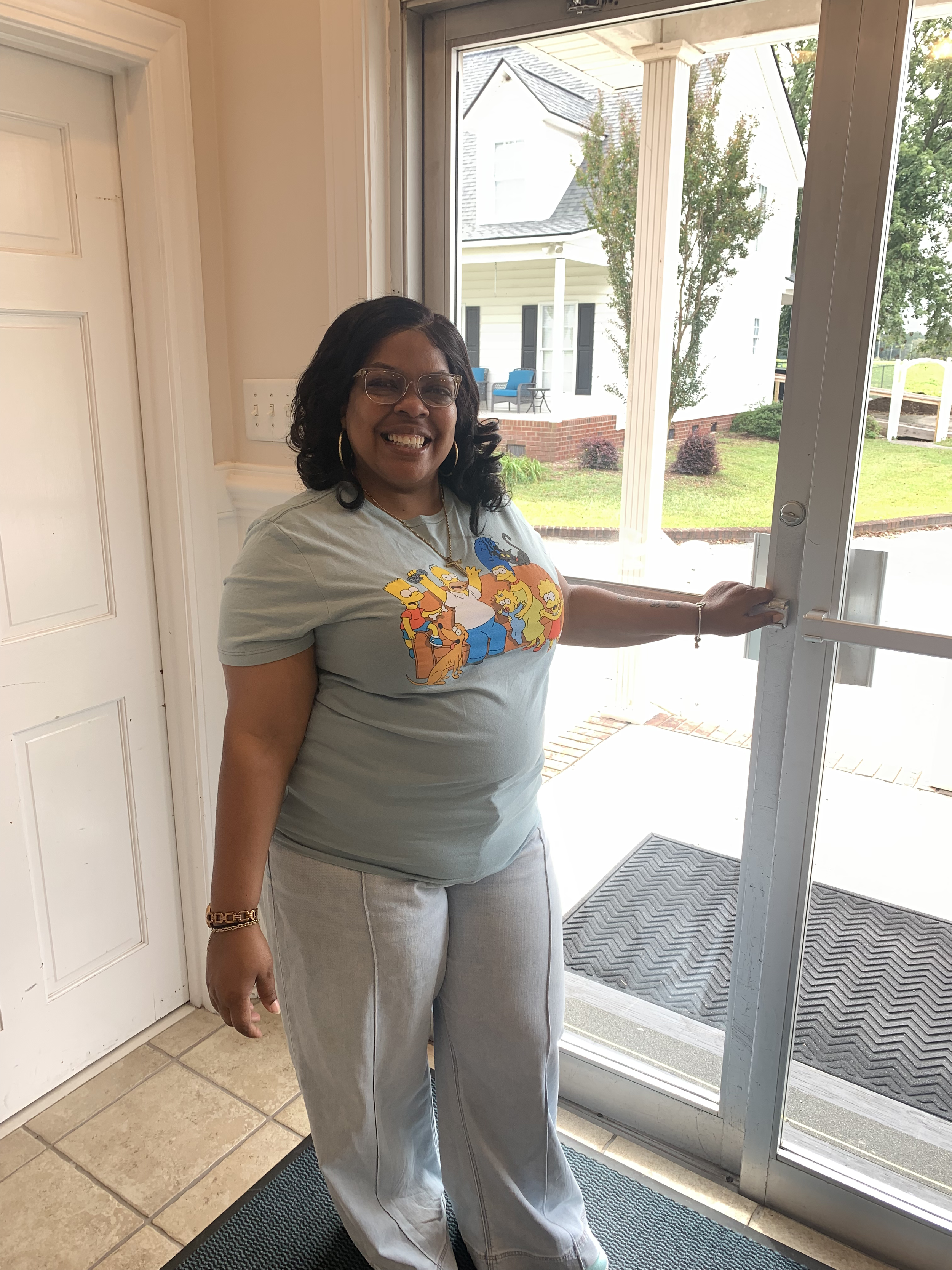 A smiling woman of Lion of Judah Community Church wearing glasses and a graphic T-shirt standing near a glass door with her hand on the door handle. 