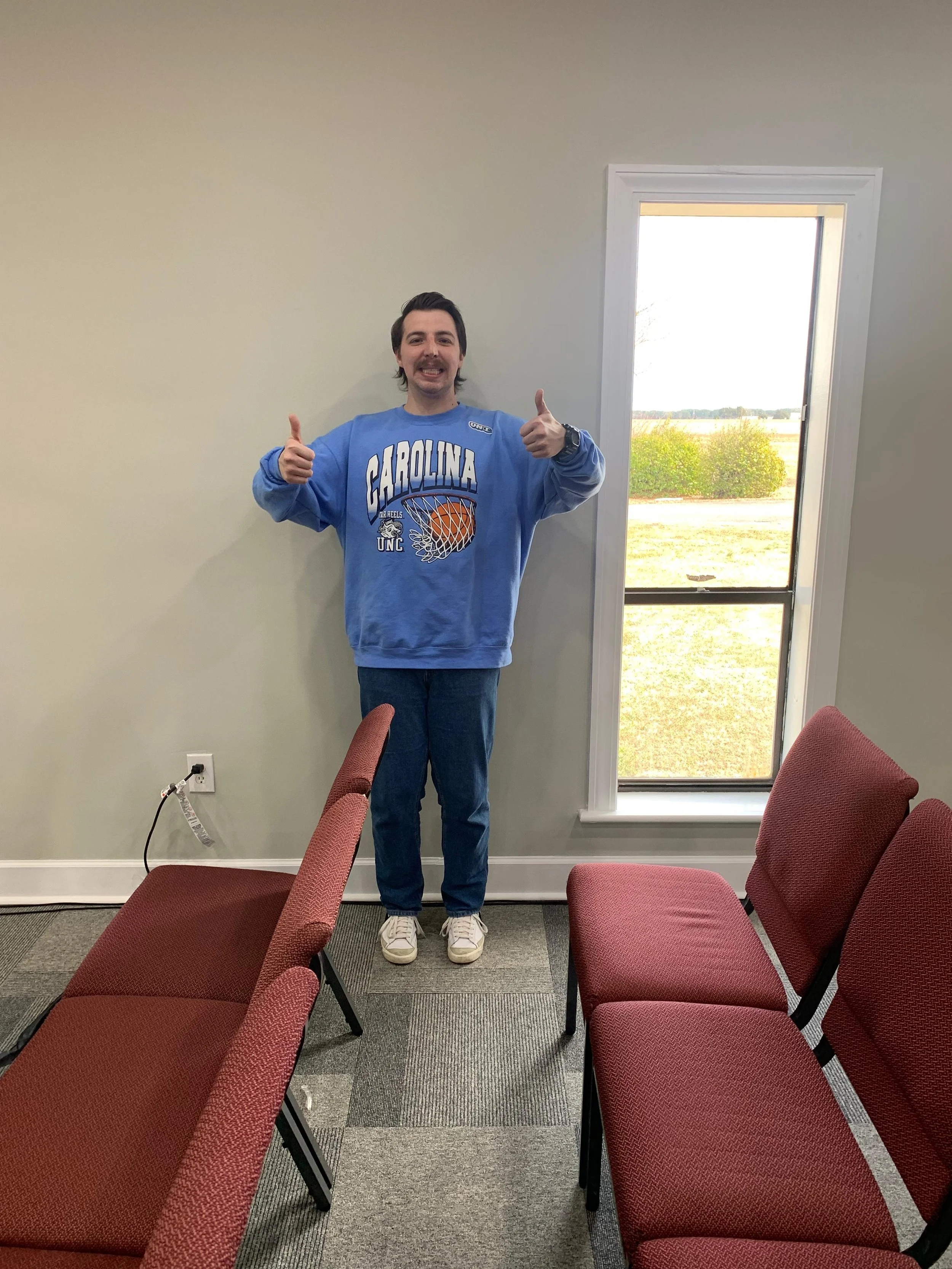 A young man with dark hair, a beard, wearing a blue Carolina basketball sweatshirt, jeans, and white sneakers, standing inside a room with beige walls and a large window showing an outdoor landscape. He is smiling and giving two thumbs up.