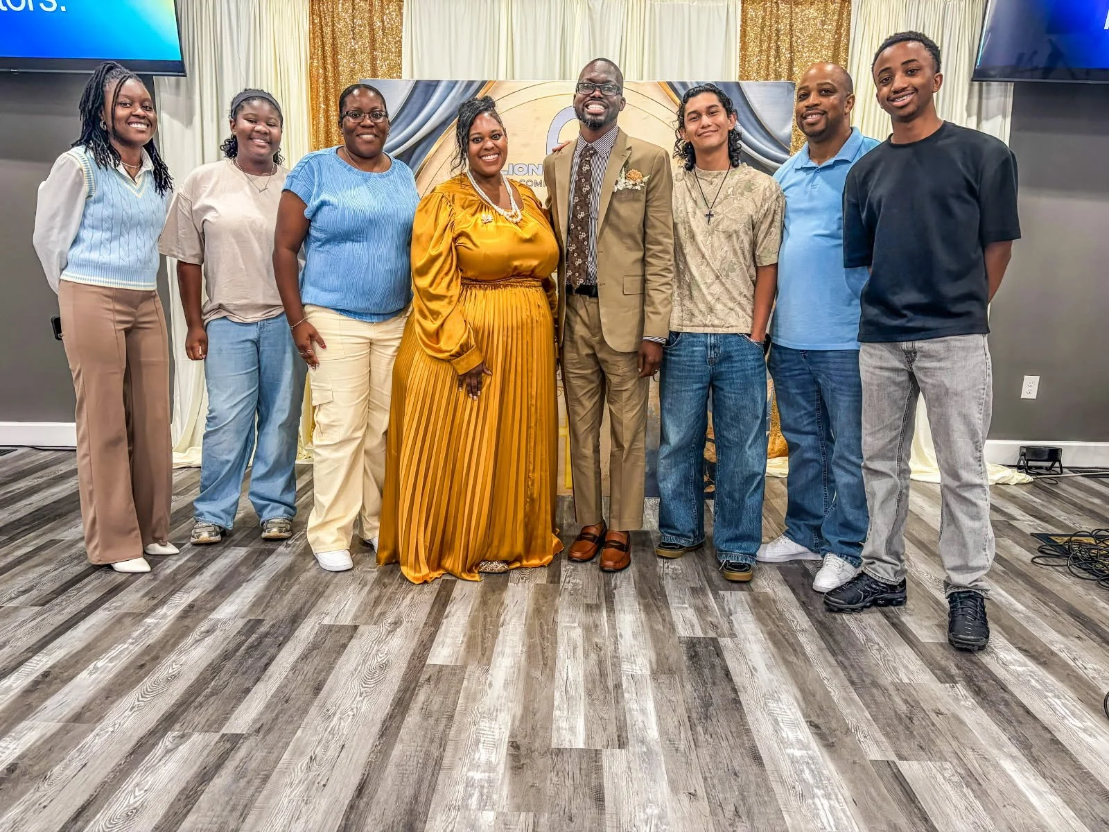 Group of nine people standing in a line indoors, posing for a photo. They are dressed in casual and semi-formal attire, smiling. Behind them is a backdrop with curtains and part of a sign or banner. The setting appears to be a celebration or event.