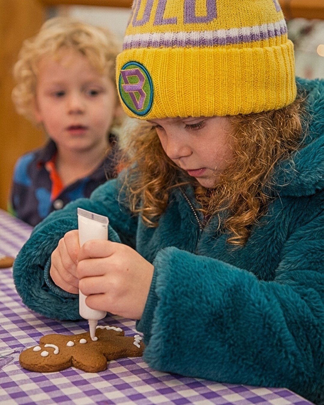 A girl wearing a yellow knit beanie called Bold Whisper from bold kidswear with purple and green details is decorating a gingerbread cookie with white icing while a boy looks on in the background.