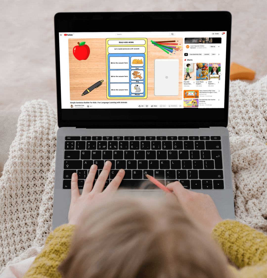 A child using a laptop on a soft, knitted blanket, watching a Bold Kids Club YouTube video about simple sentence building for kids with animals, with colorful educational graphics on the screen.
