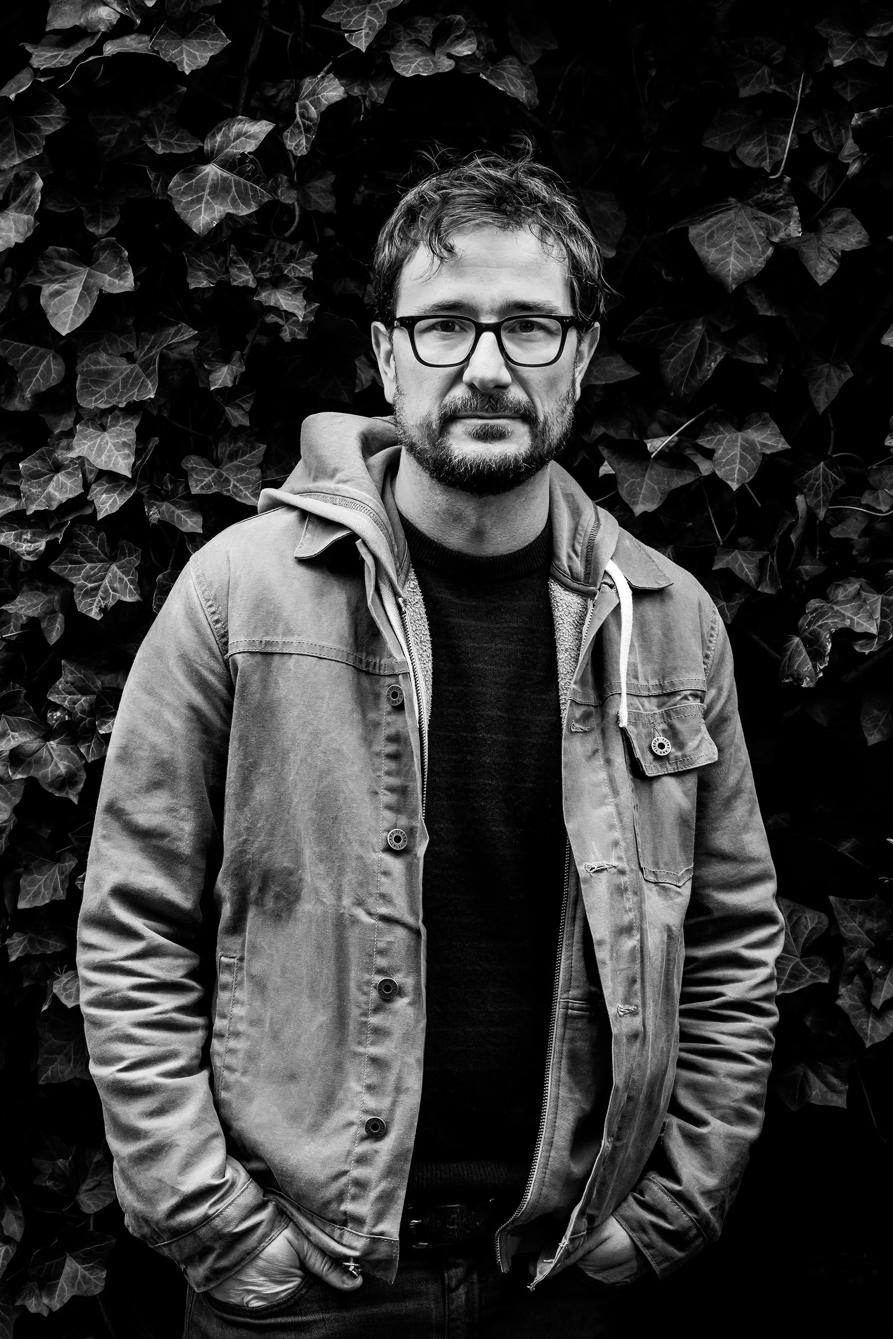 Black-and-white portrait of a bearded man wearing glasses and a jacket in front of ivy leaves