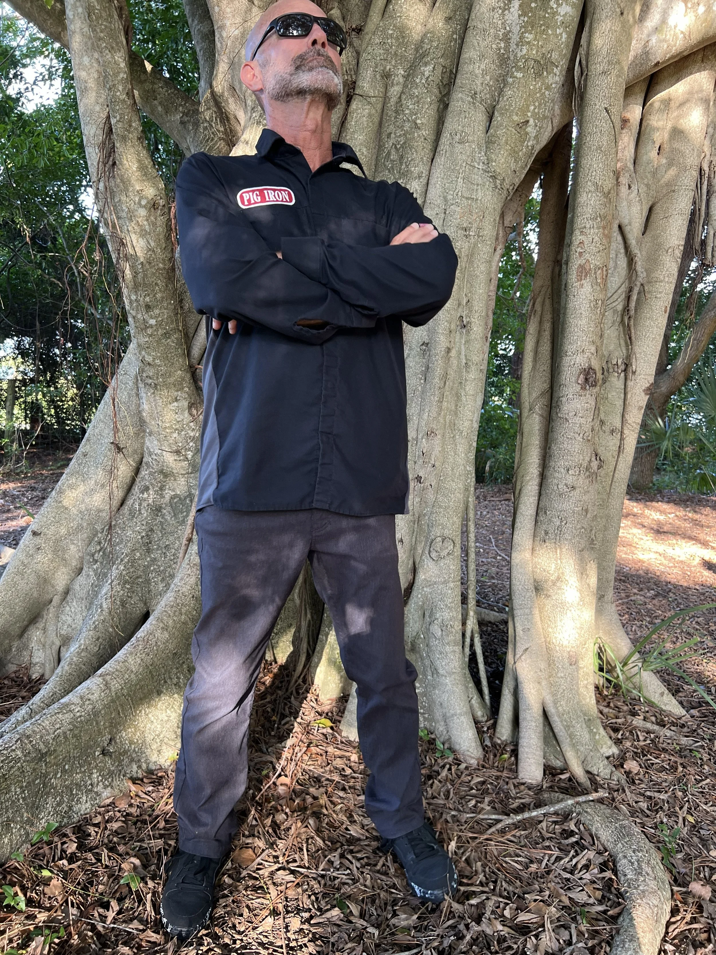 A man with sunglasses, a gray beard, and a goatee, dressed in a black jumpsuit with a Patch that says 'Pig Iron,' stands with arms crossed in front of a large tree with thick roots and branches. The setting appears to be outdoors in a wooded area with fallen leaves and greenery.