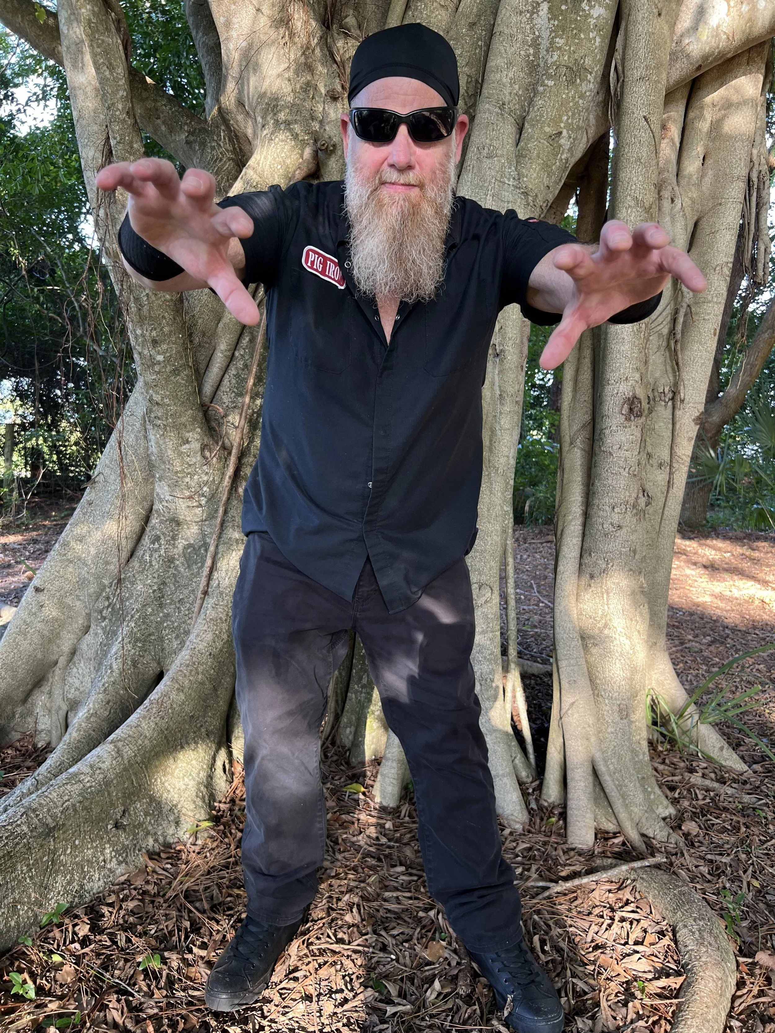 A man with a long beard wearing sunglasses, a black head covering, and black clothing stands in front of a large tree with many roots and branches, extending his arms forward. The man is outdoors in a wooded area with leaves on the ground.