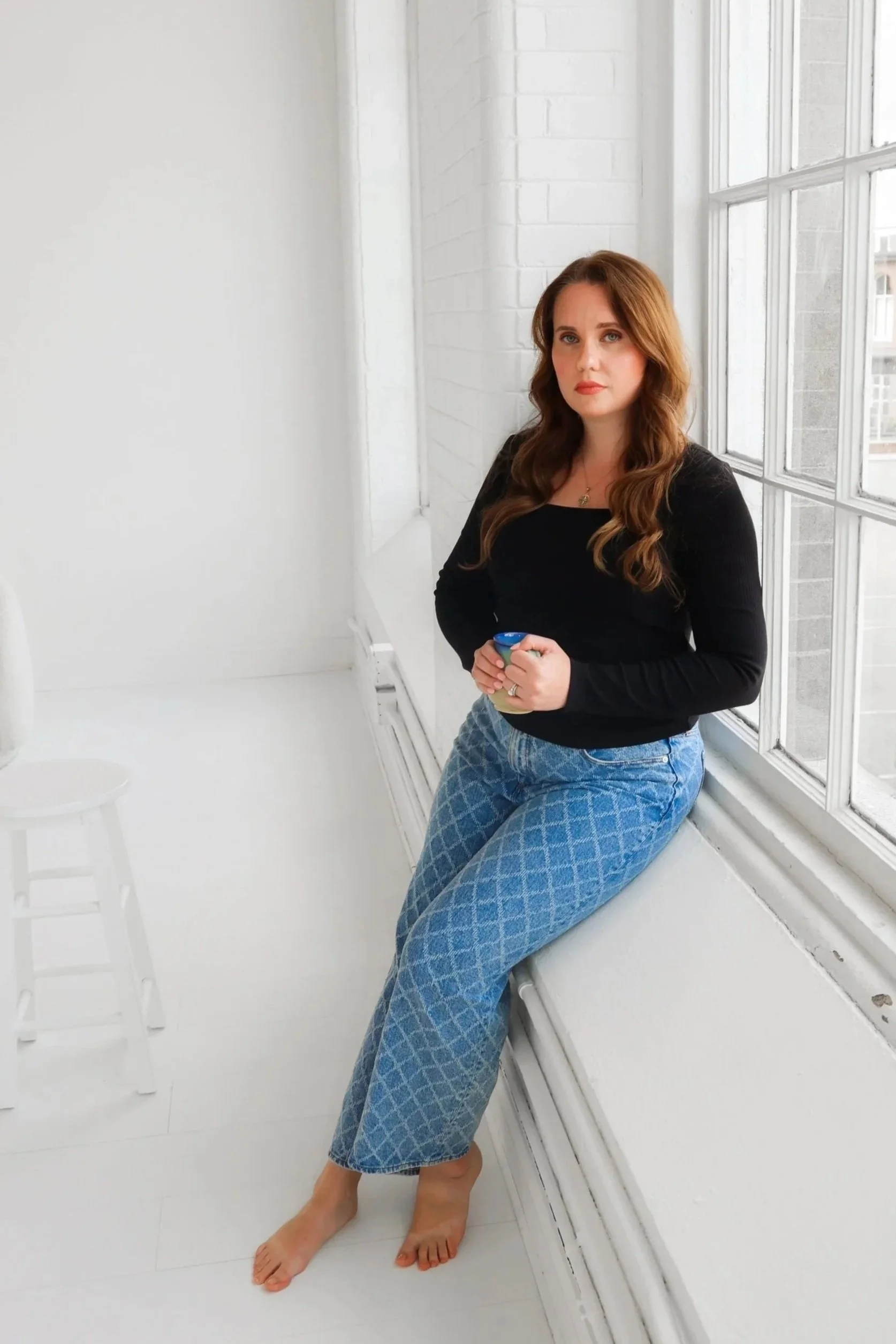 A woman with long, wavy auburn hair, wearing a black long-sleeve shirt and blue patterned pants, is sitting on a white windowsill in a bright room with white walls and large windows, holding a mug with both hands.