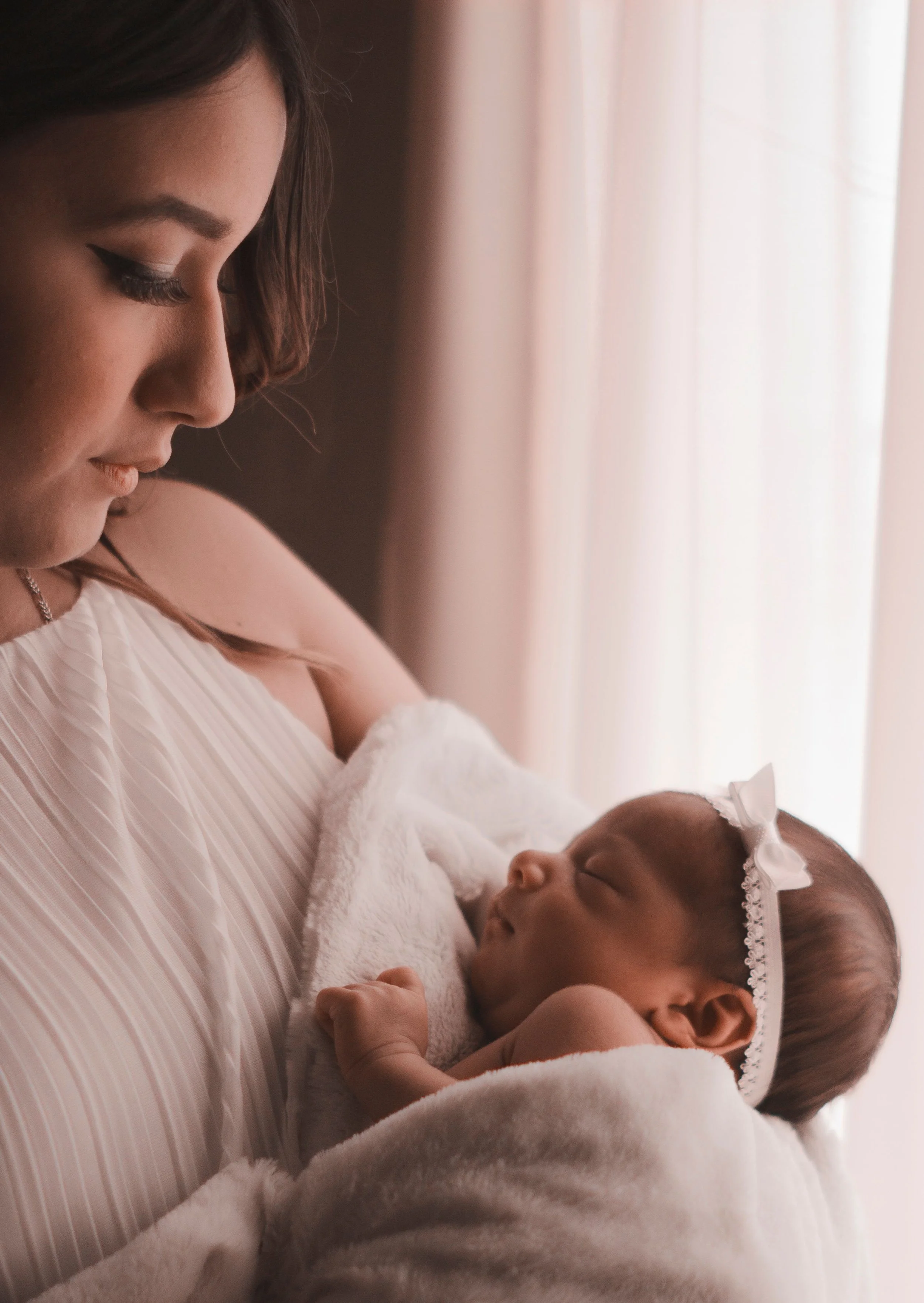 A woman holding a sleeping baby wrapped in a white towel near a window with light curtains.