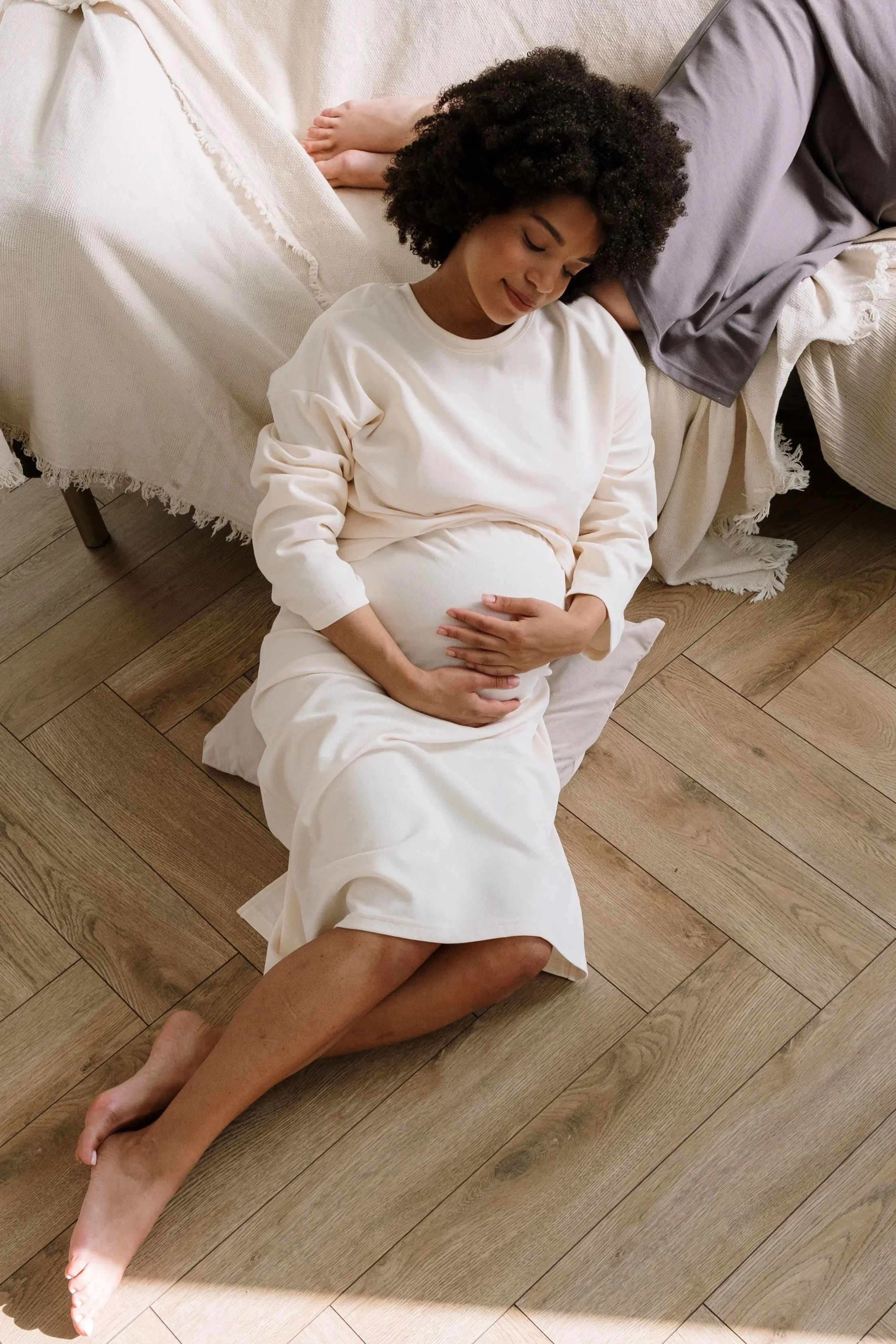 A pregnant woman sitting on the floor with her eyes closed, leaning back against a bed with a gray blanket, and resting her hand on her belly, while holding a man's hand on her legs.