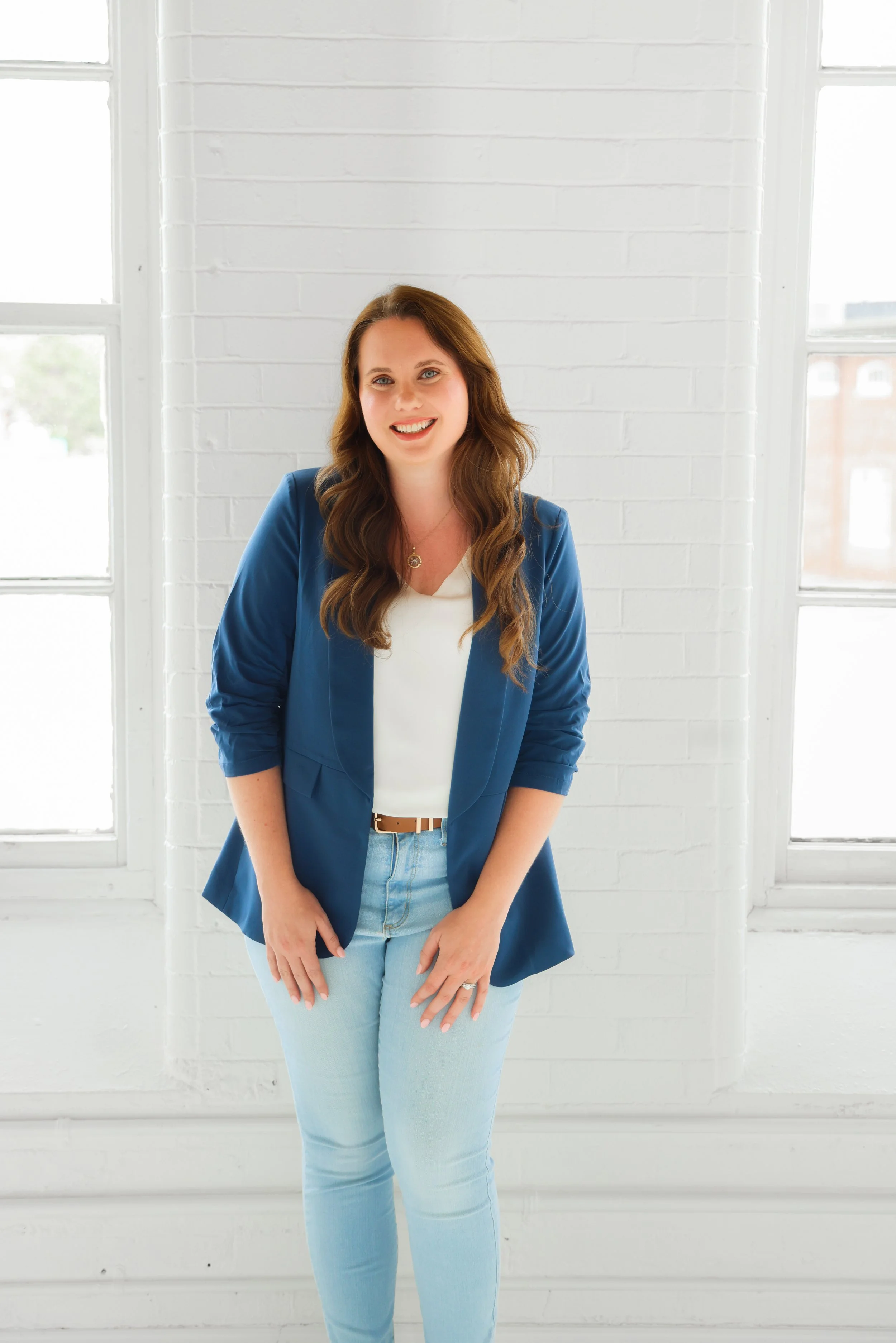 A woman with long brown hair smiling, wearing a blue blazer, white top, and light blue jeans, standing in front of a white brick wall with large windows on either side, bright natural light.