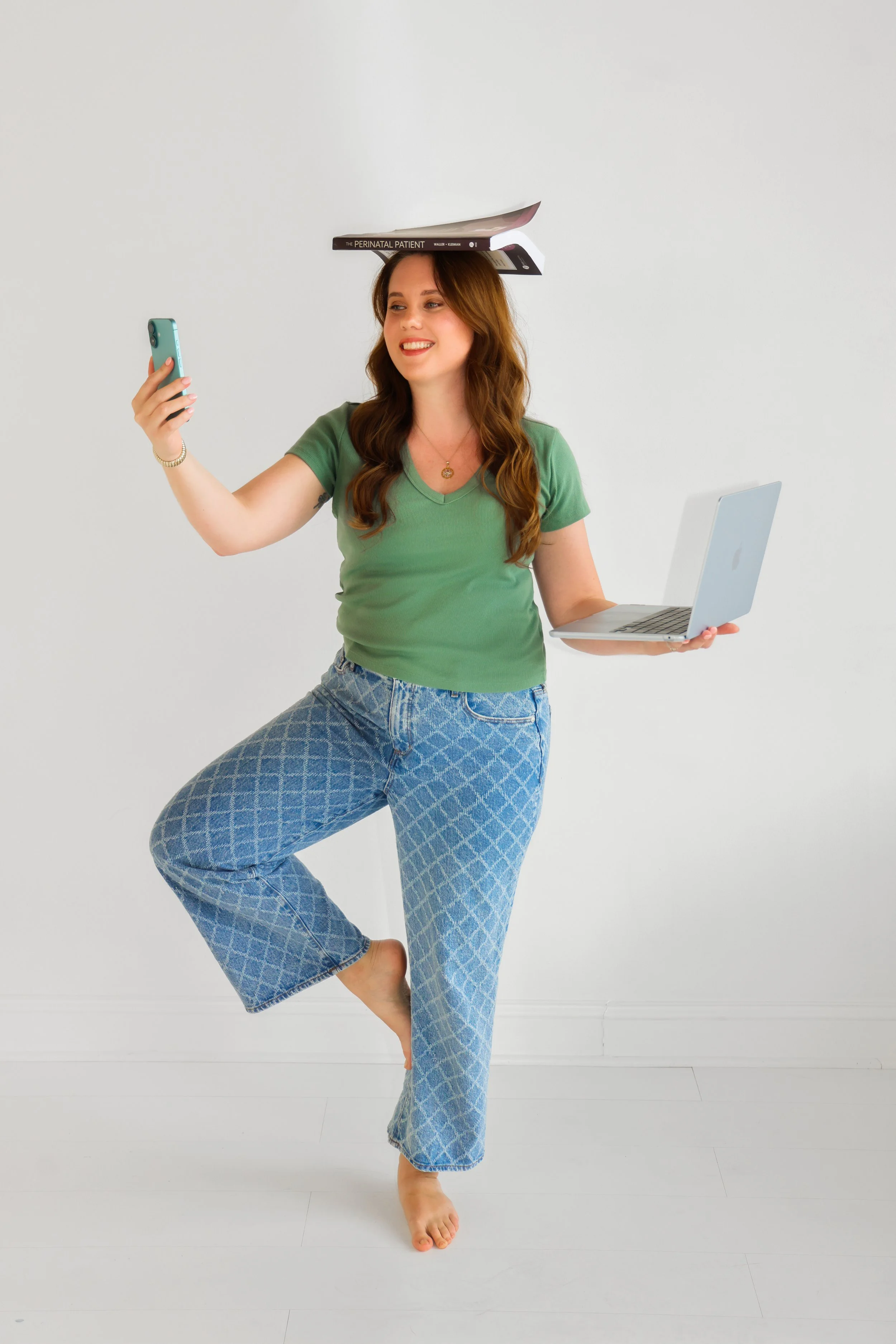 A woman in a green t-shirt and patterned jeans balancing on one foot, holding a laptop in one hand and a phone in the other, with books on her head.